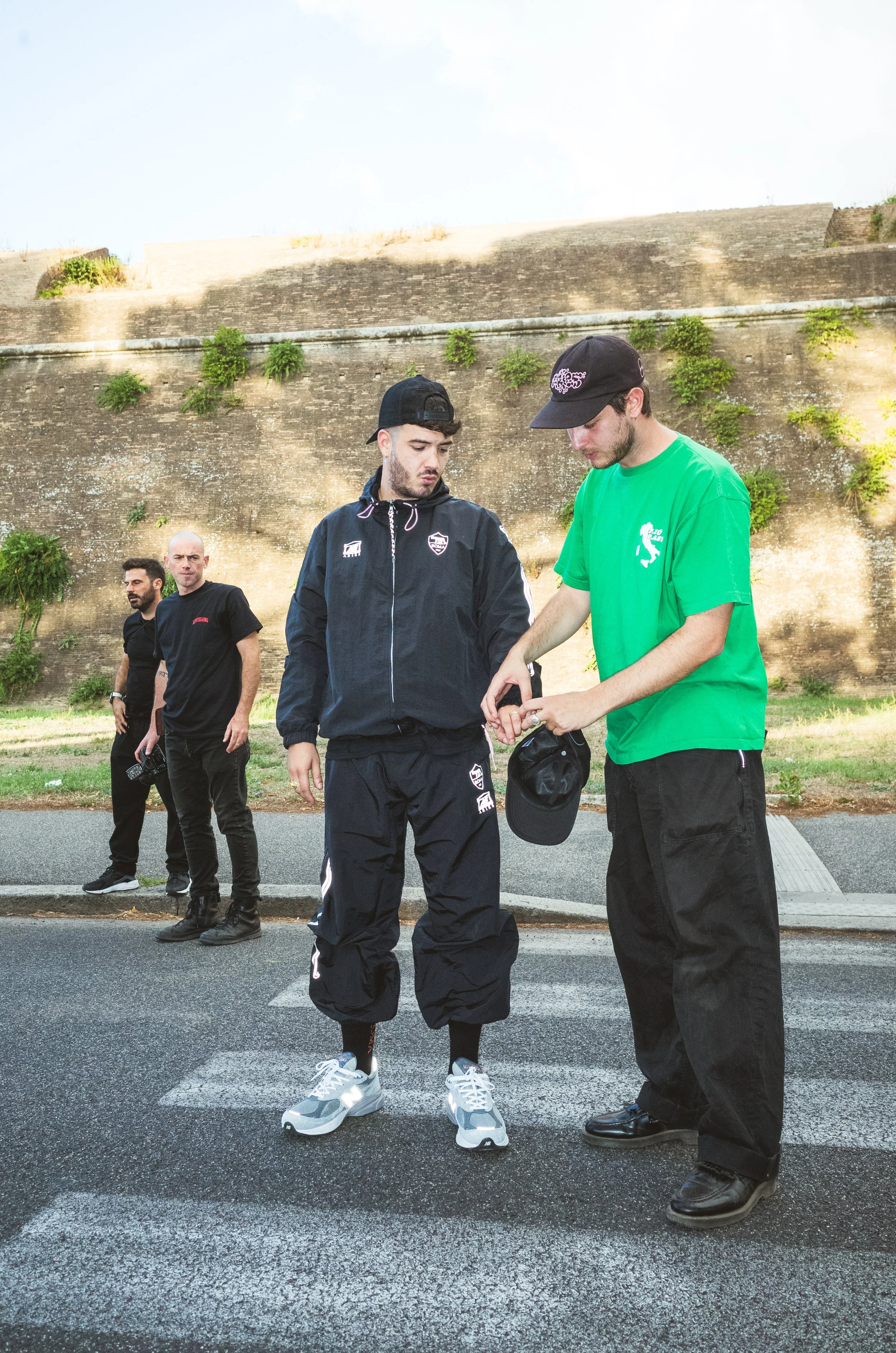 Two young men are standing on a crosswalk, one in black athletic clothing and the other in a green T-shirt, looking at something in the other man's hand. Two other men are standing behind them on the street curb, watching. A large old stone wall with