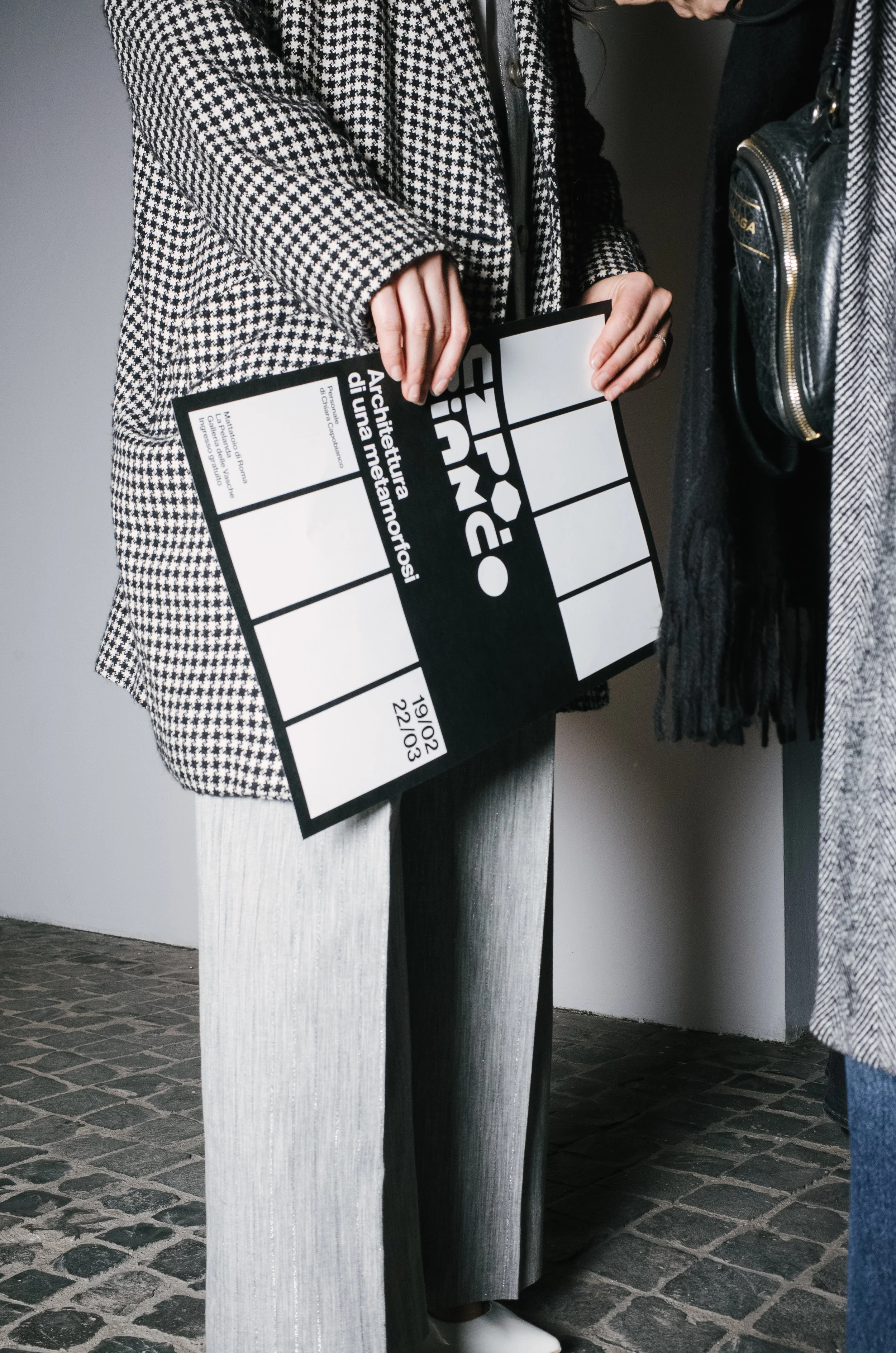 Close-up of a person holding a film slate or clapperboard with people around, wearing stylish clothing, standing on a cobblestone floor.