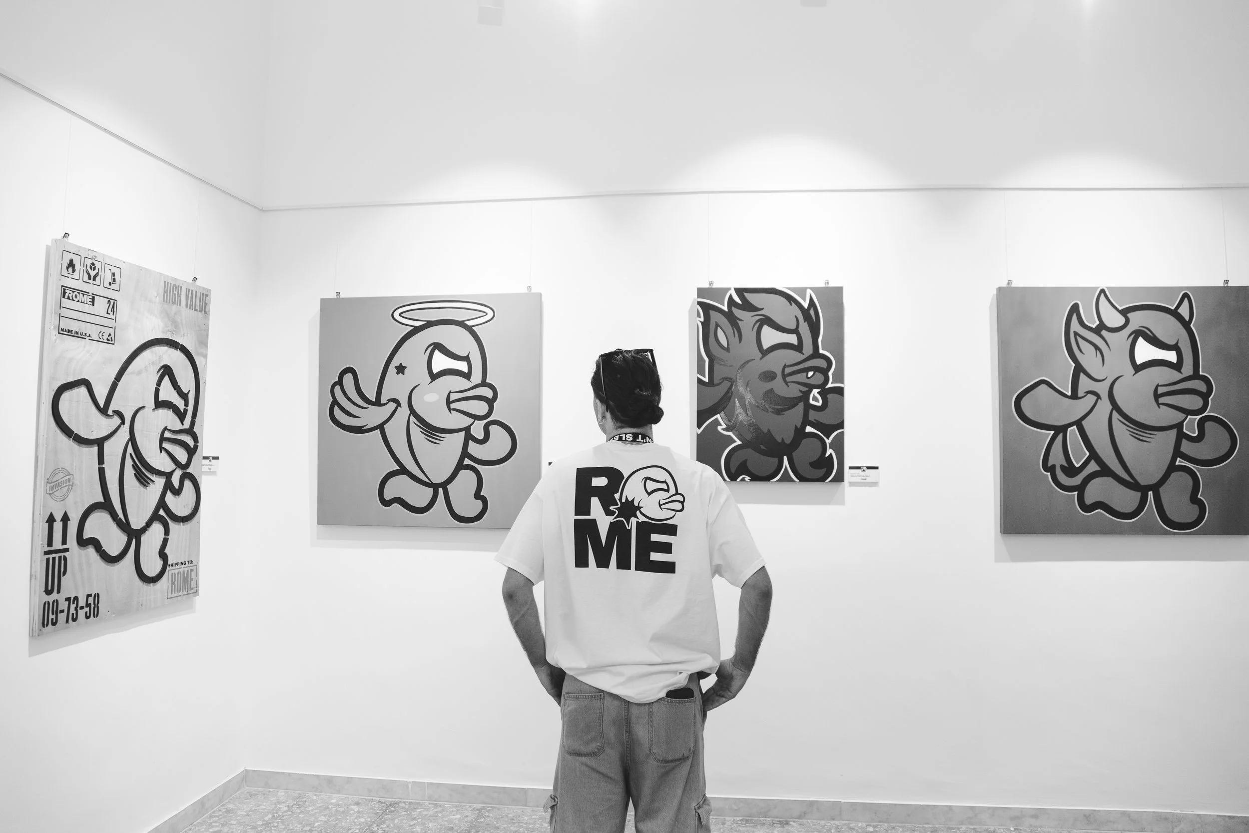 Person wearing a T-shirt with the word 'ROME' and a cartoon of a fish with a tongue out, viewing cartoon fish artwork in a gallery