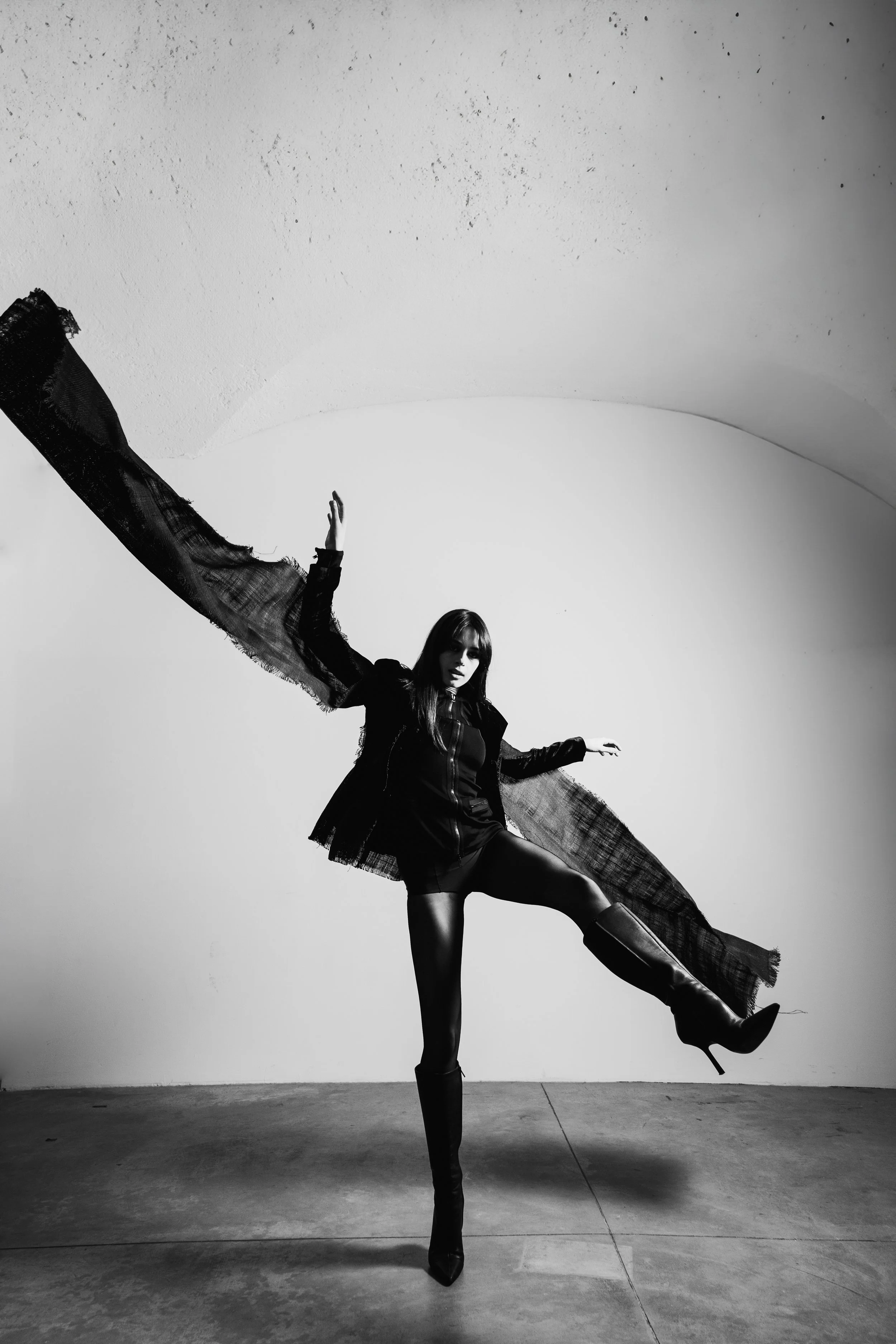 Black and white photo of a woman posing with one leg raised and arms outstretched, wearing high boots and a flowing coat.