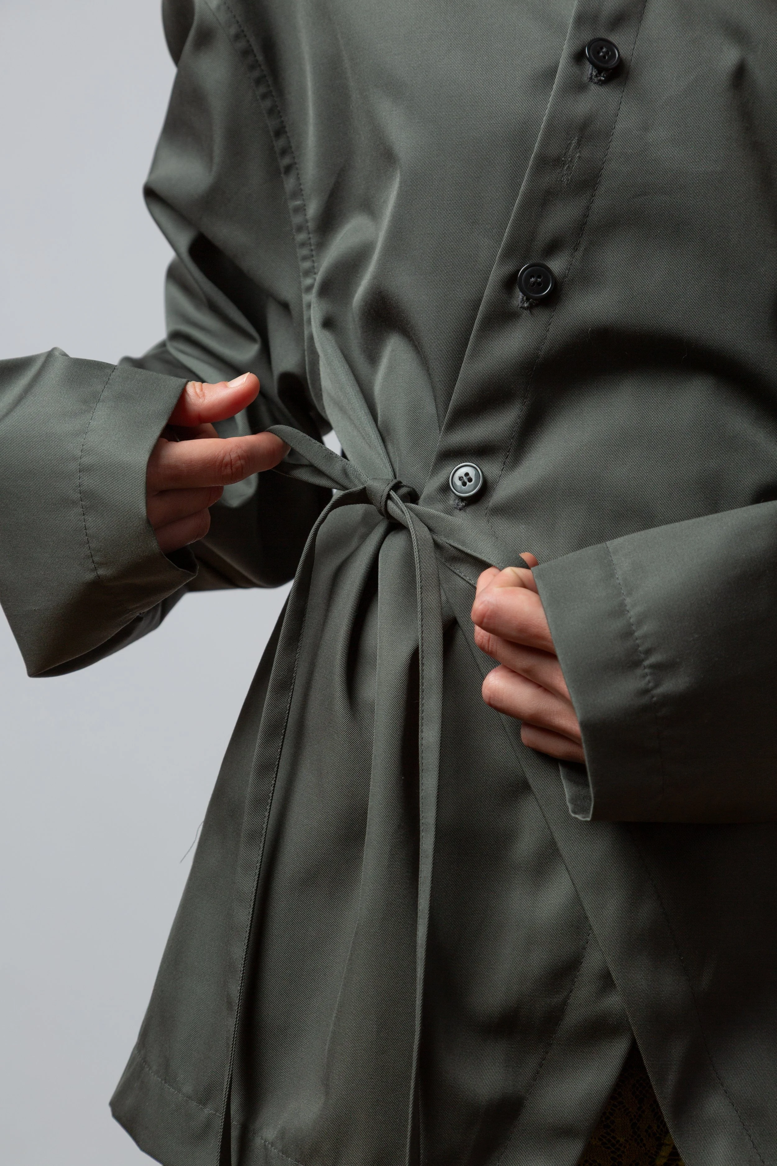 Person tying the belt of a gray coat with buttons.