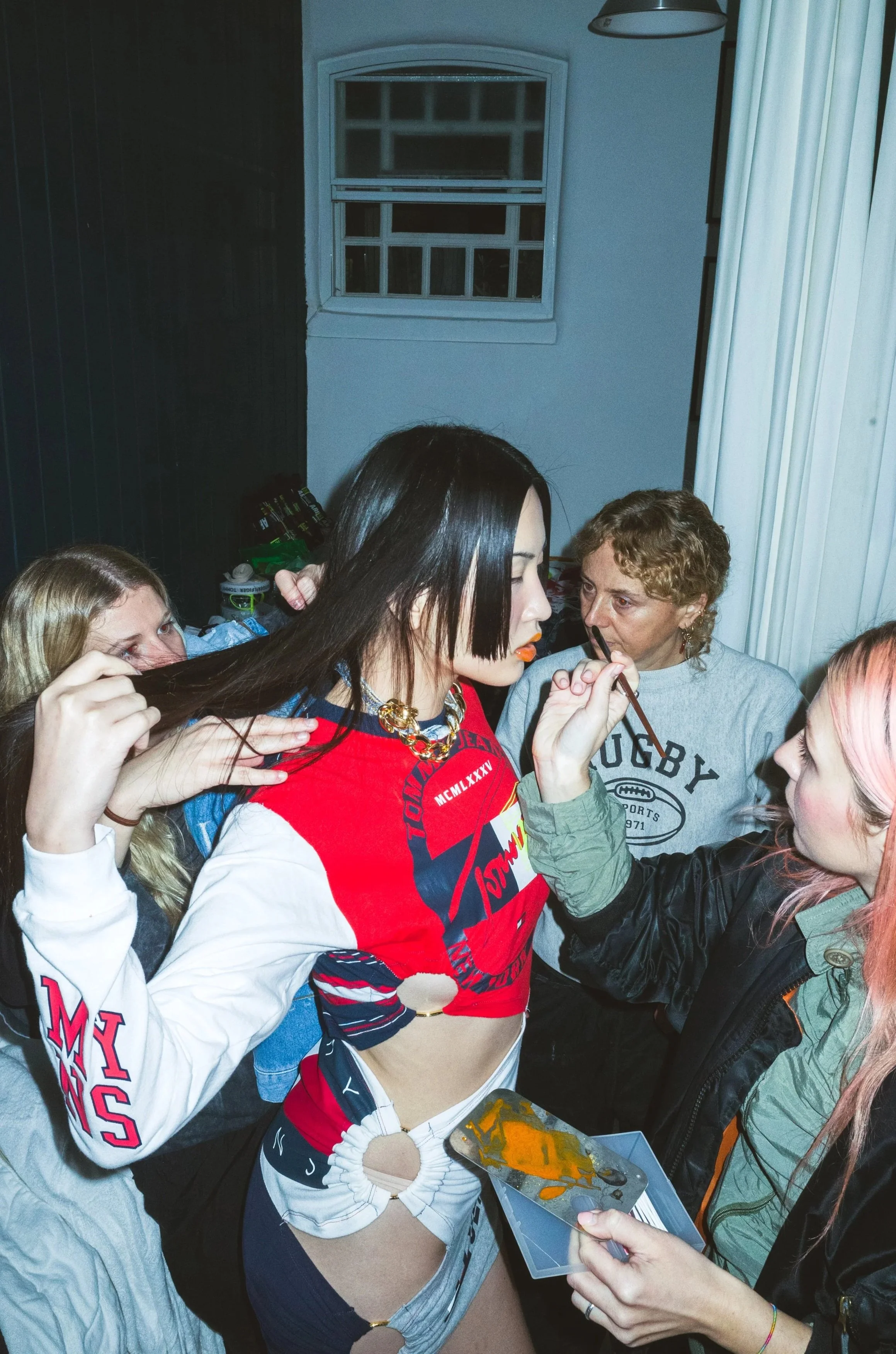 A young woman with black hair and colorful makeup being painted by a girl with pink hair during a social gathering, with other women around them in a room with a window and curtains.