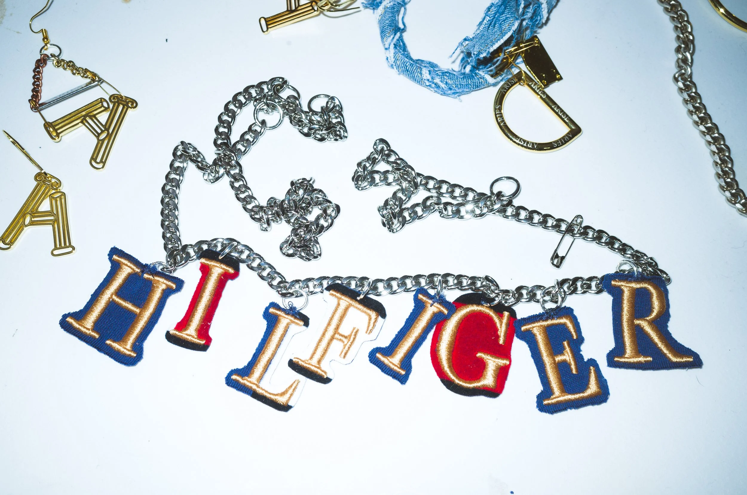 A silver chain necklace with colorful embroidered letters spelling 'HIGHER' and various scattered jewelry pieces on a white surface.