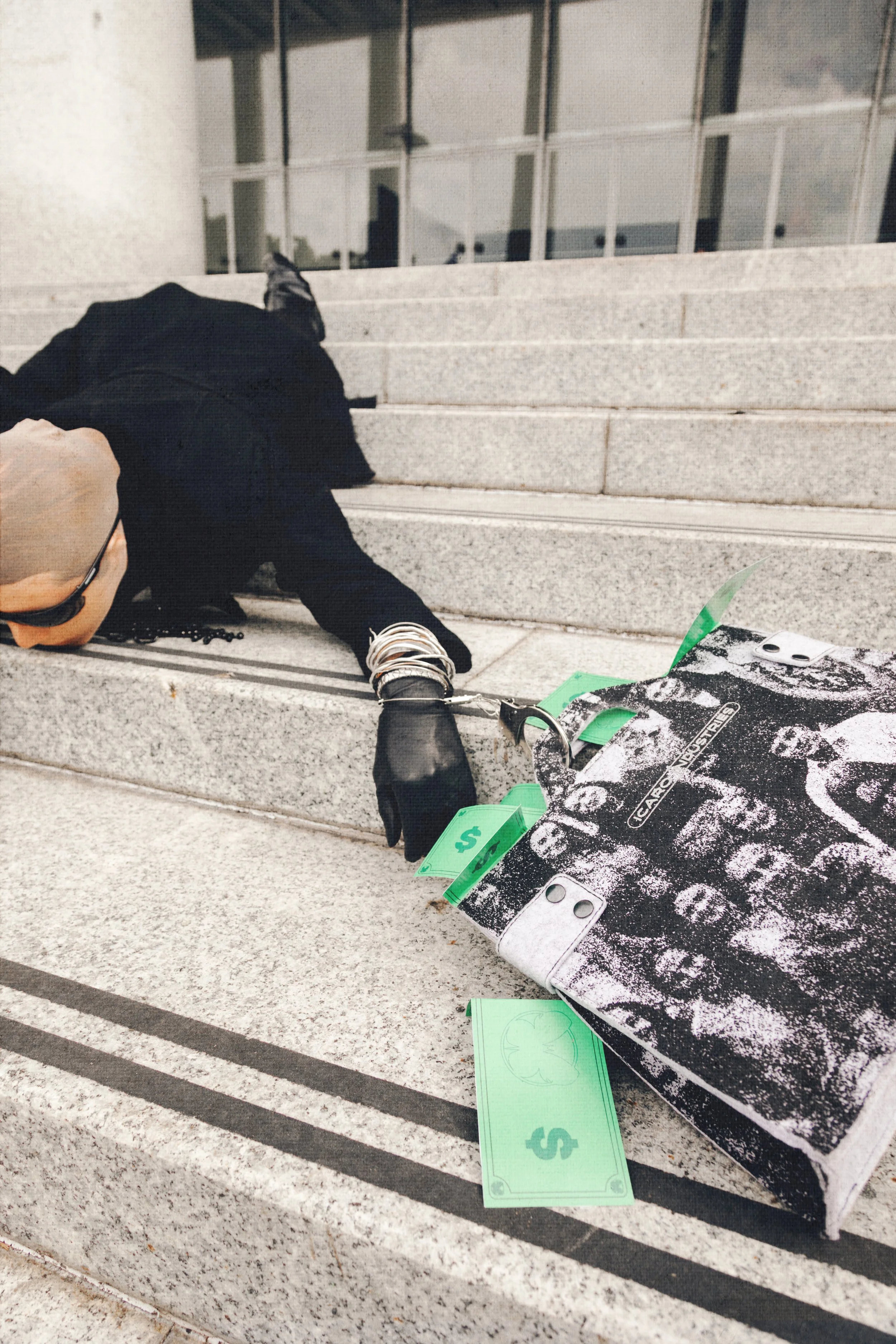 A person lying on granite steps, wearing sunglasses and a hoodie, with a skateboard and fake money around them.