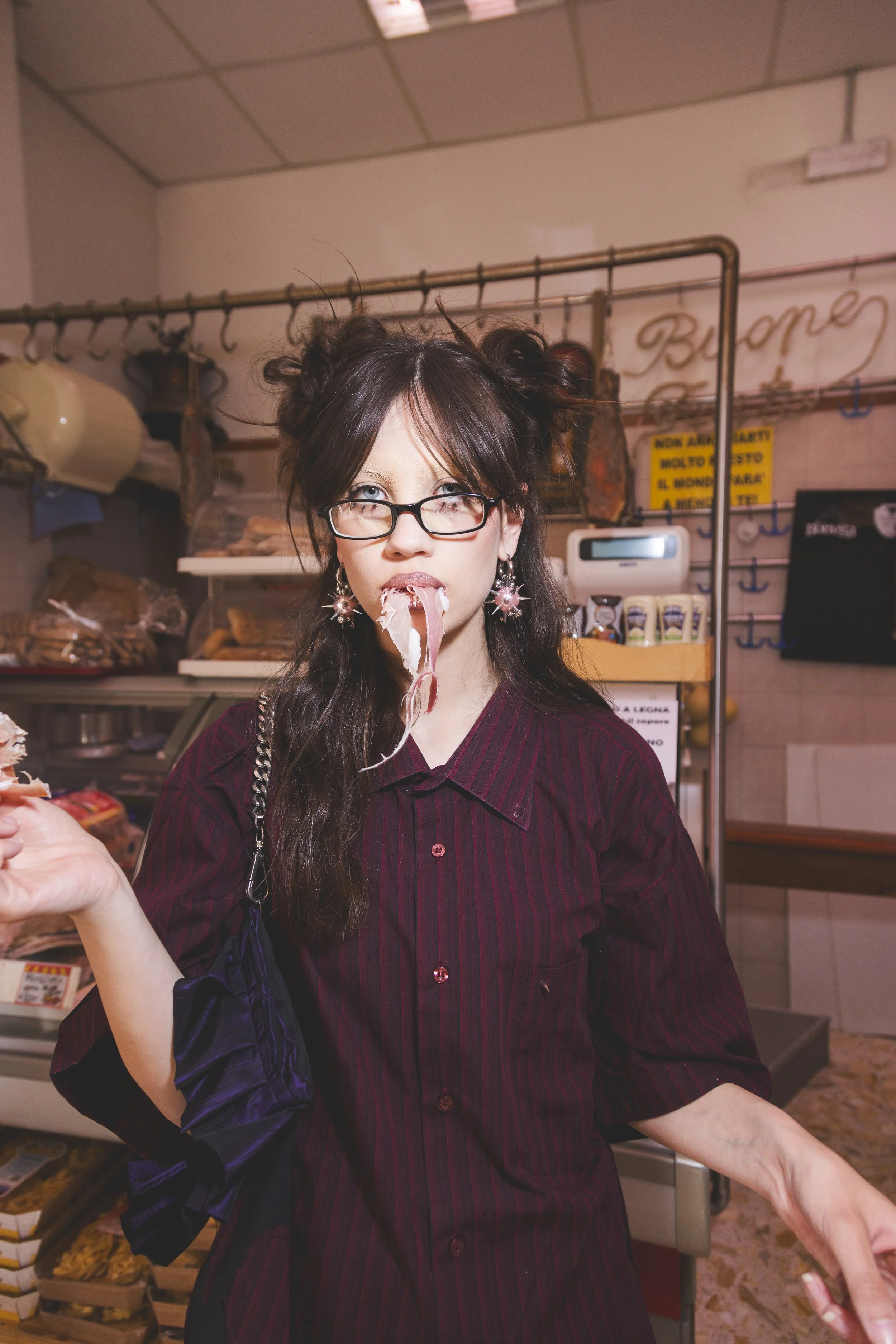 A woman with glasses and dark hair styled in buns is in a store, eating a piece of meat with a playful and surprised expression.