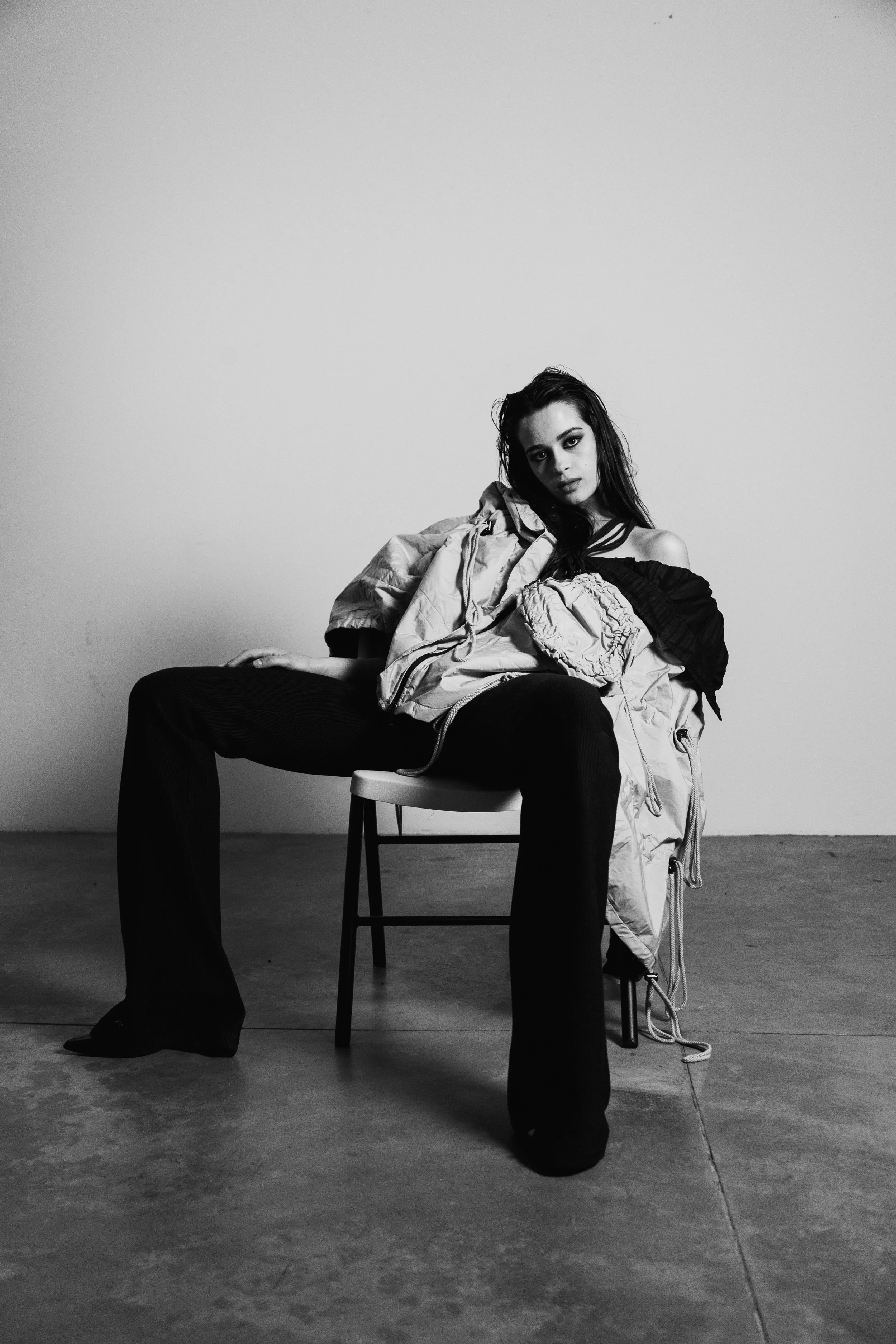 A black-and-white photo of a young woman sitting on a chair with her legs spread apart and leaning to one side. She is wearing a loose jacket and has dark, wet-looking hair, with a serious expression on her face. The setting is a plain, empty room wi