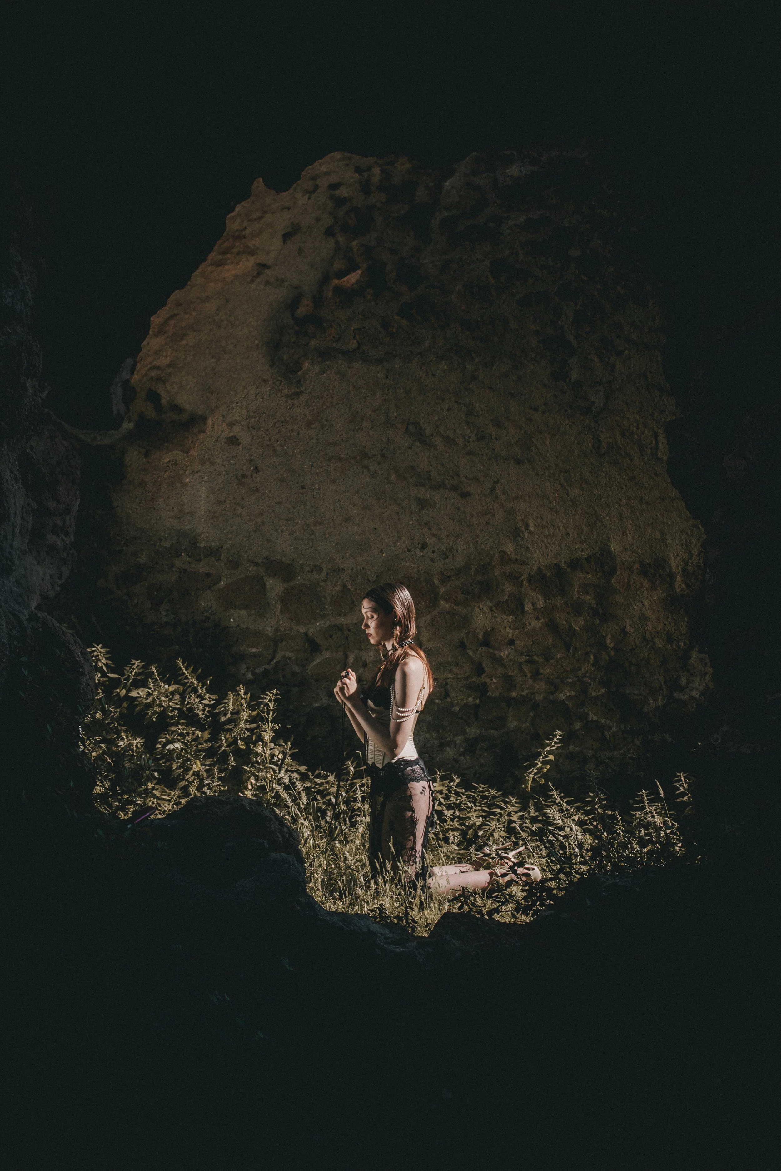 A woman kneeling among rocks and plants in a natural setting, gazing downward, dark background with large rock formation.