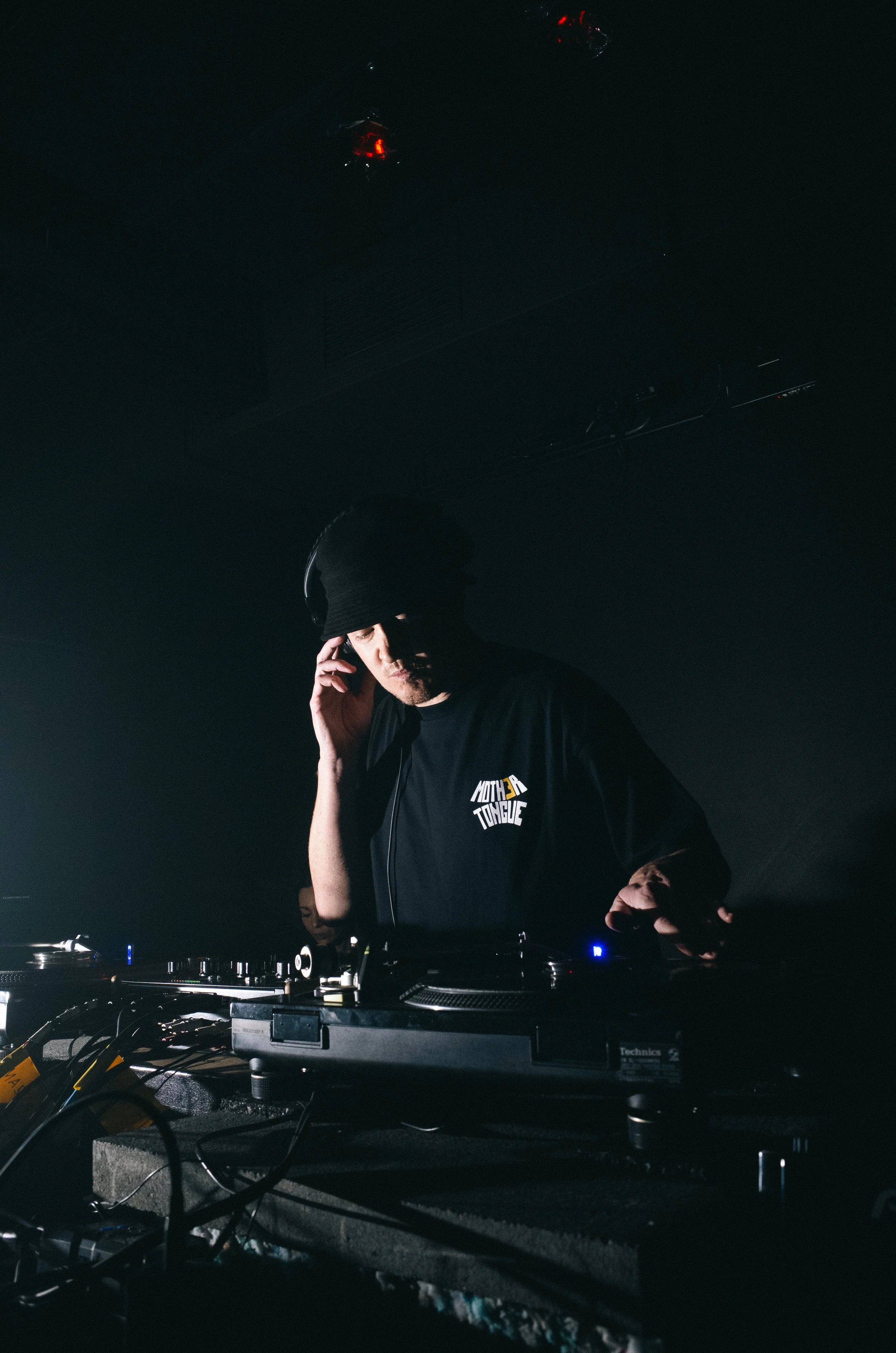 DJ wearing black clothing and a beanie, standing behind turntables and mixing equipment, in a dark club or studio environment.