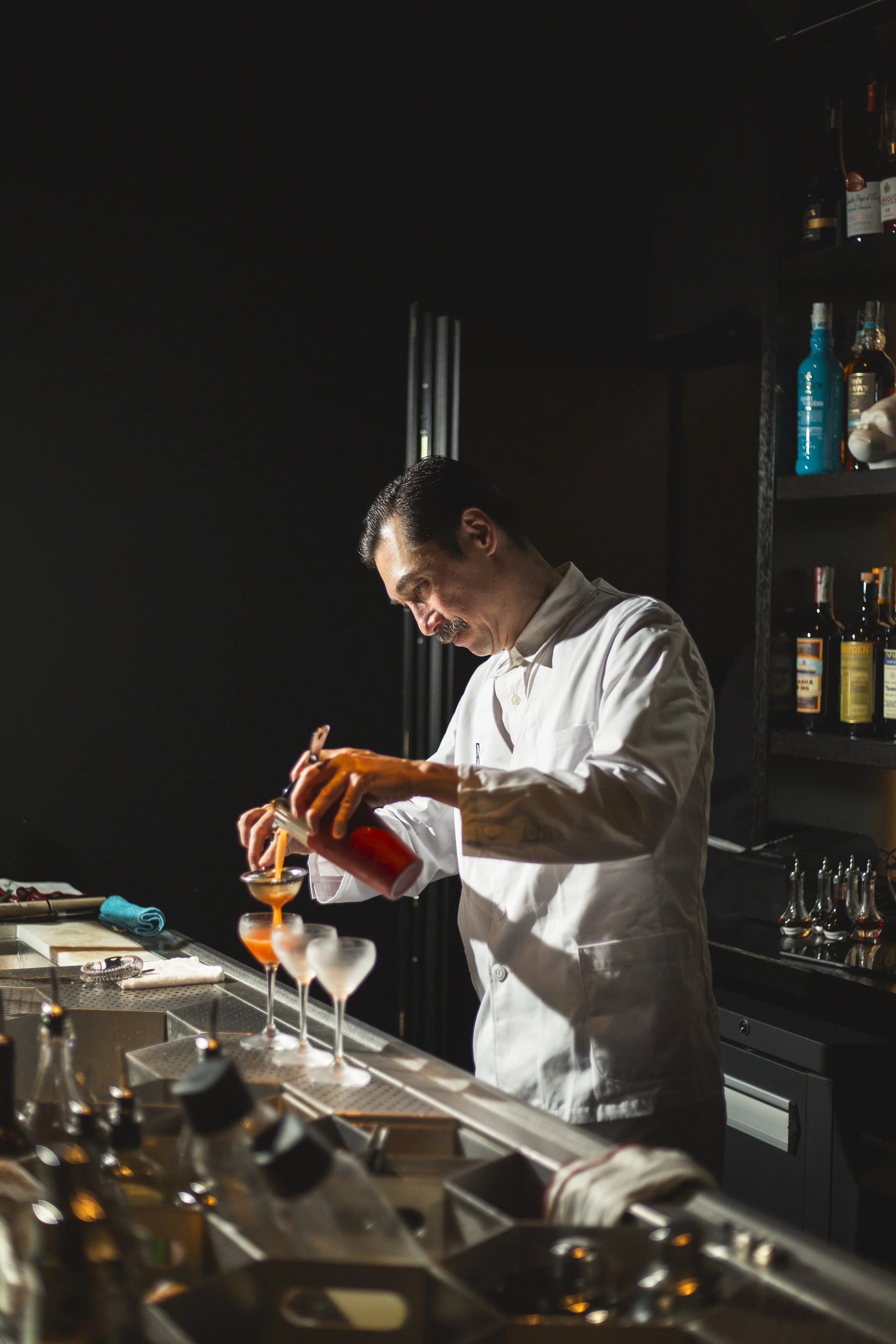 A bartender in a white coat prepares cocktails behind a bar, pouring an orange liquid into martini glasses.
