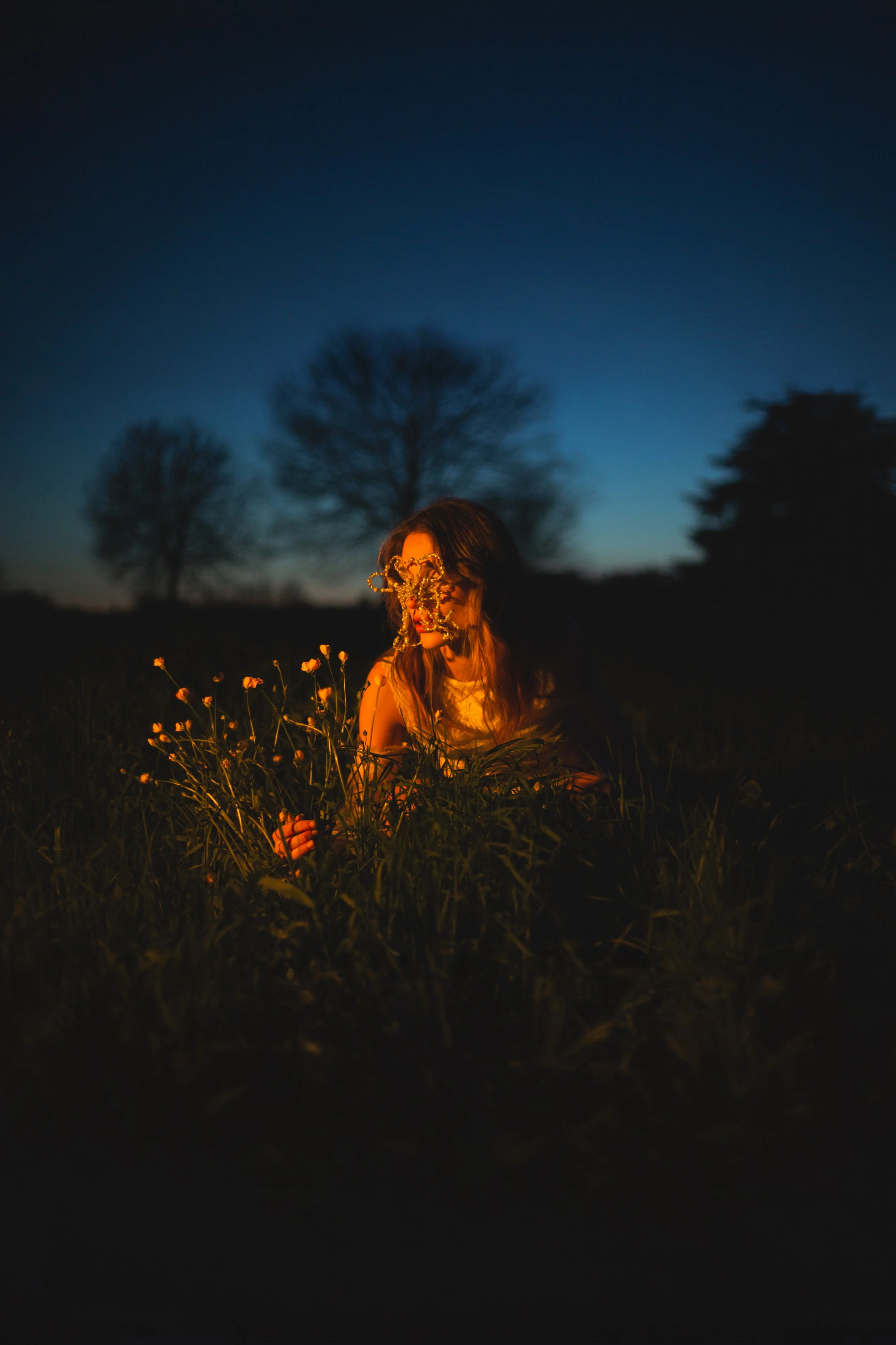 A woman sitting in a grassy field at dusk, wearing a floral mask and illuminated by warm light, with trees and a darkening sky in the background.