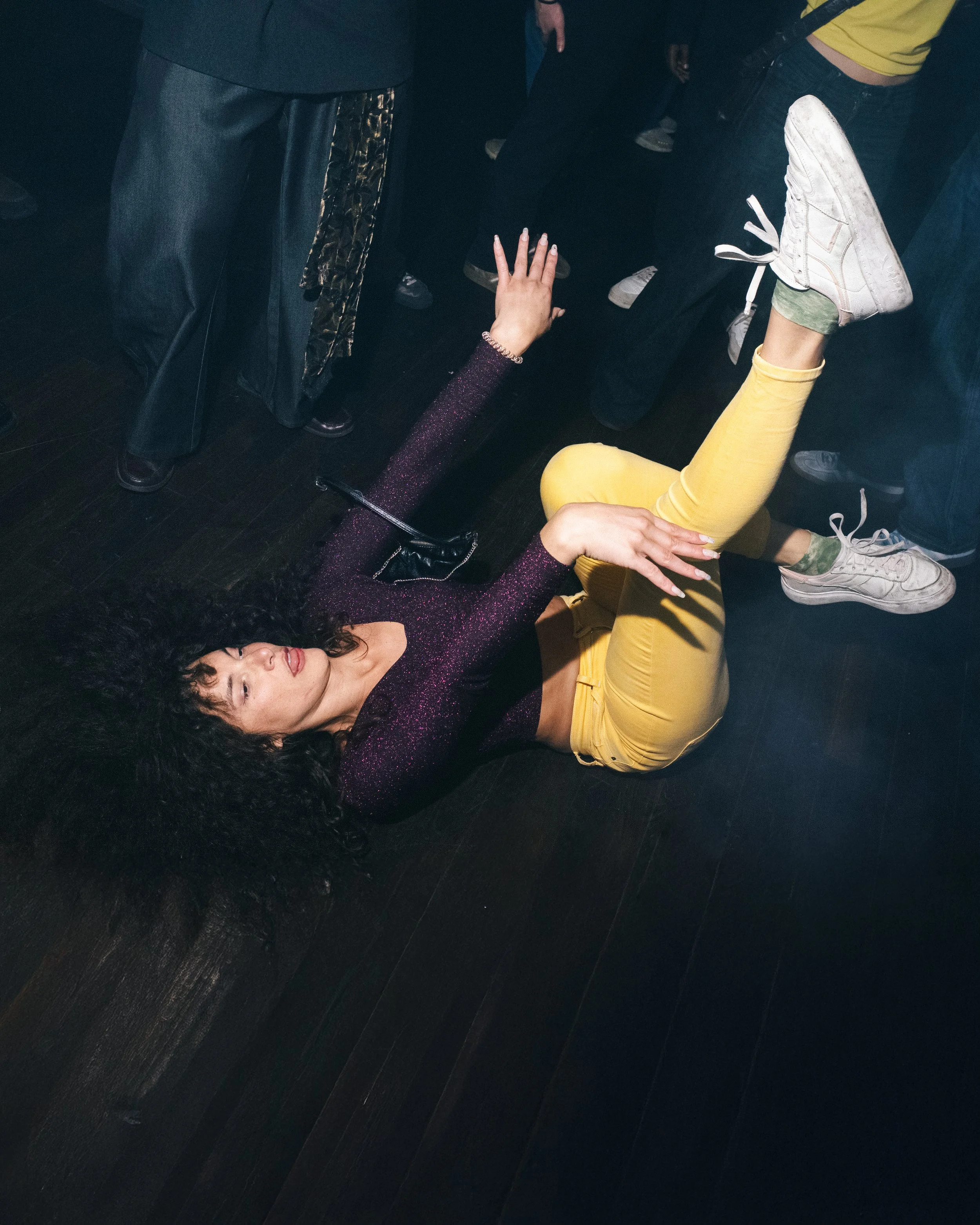 A woman with curly black hair lies on the dark floor, reaching upward with her left hand, surrounded by people at a social event.