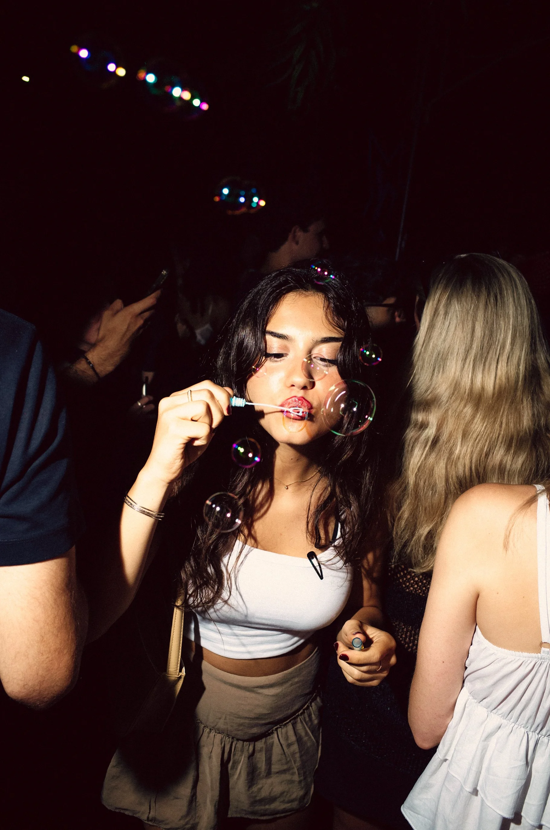 A young woman with dark wavy hair blowing bubbles at a party or club, surrounded by other people, with colorful lights in the background.