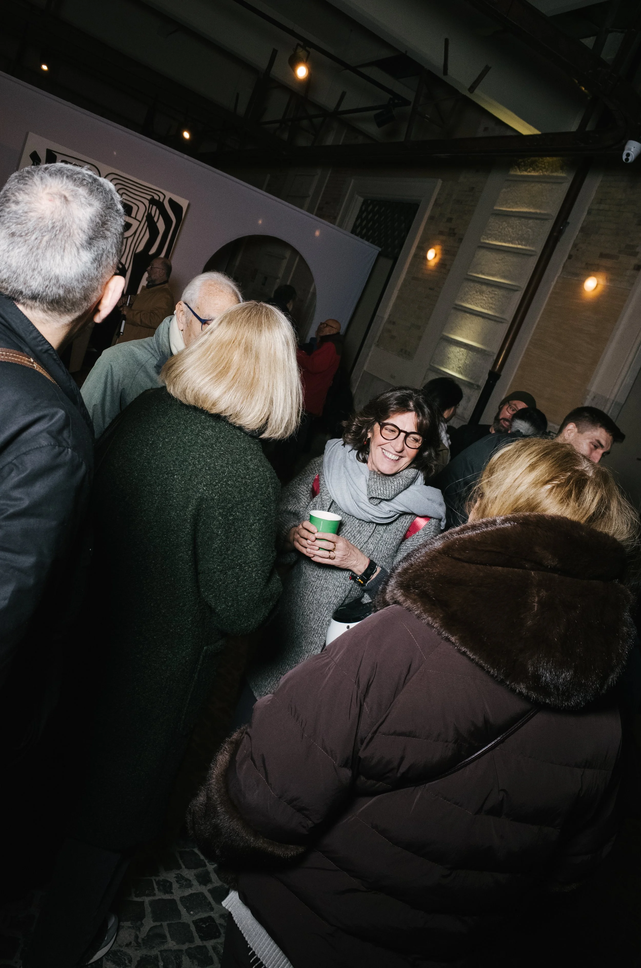 Group of people socializing at an indoor event, with a smiling woman wearing glasses holding a cup.