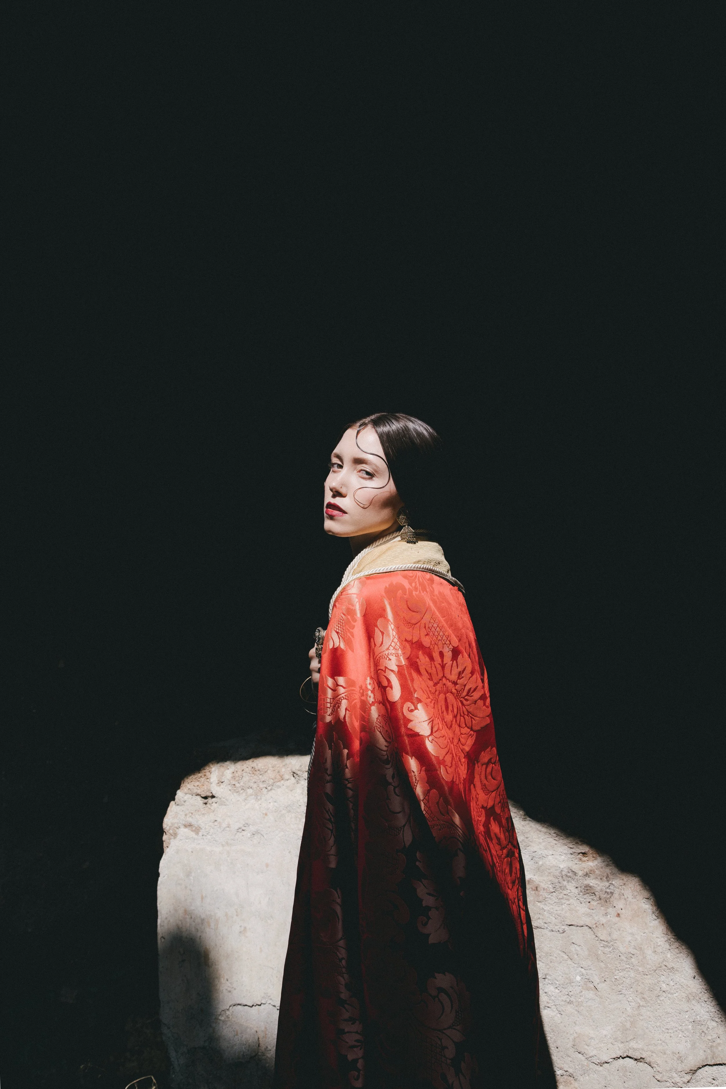 A woman with dark hair and red lipstick wearing an ornate red and gold robe, standing in front of a black background with a large rock, looking over her shoulder.