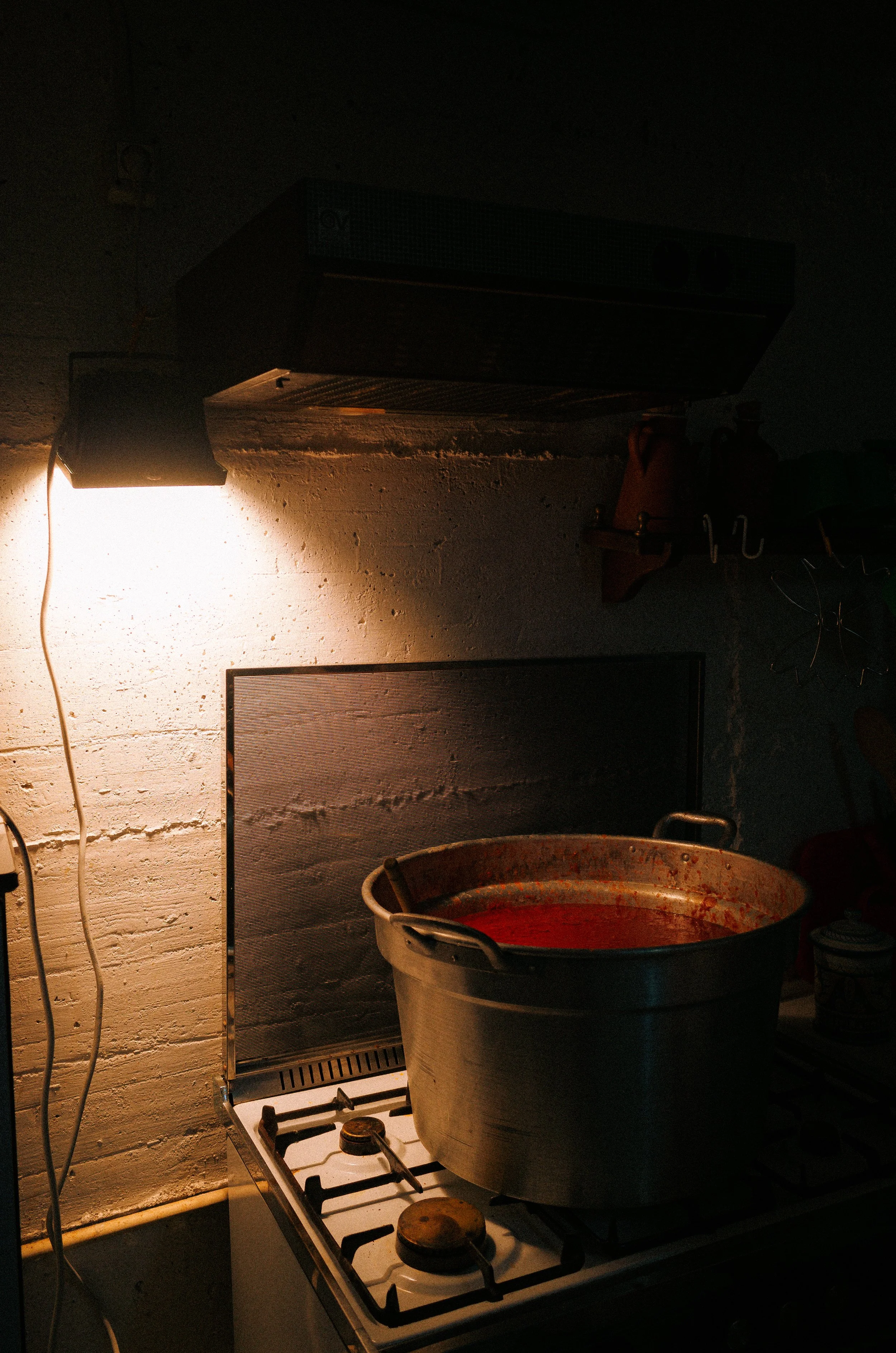 A large pot with tomato sauce on a stovetop in a dimly lit kitchen. The pot is placed on the front right burner of a gas stove, with a white brick wall and a vent hood above.