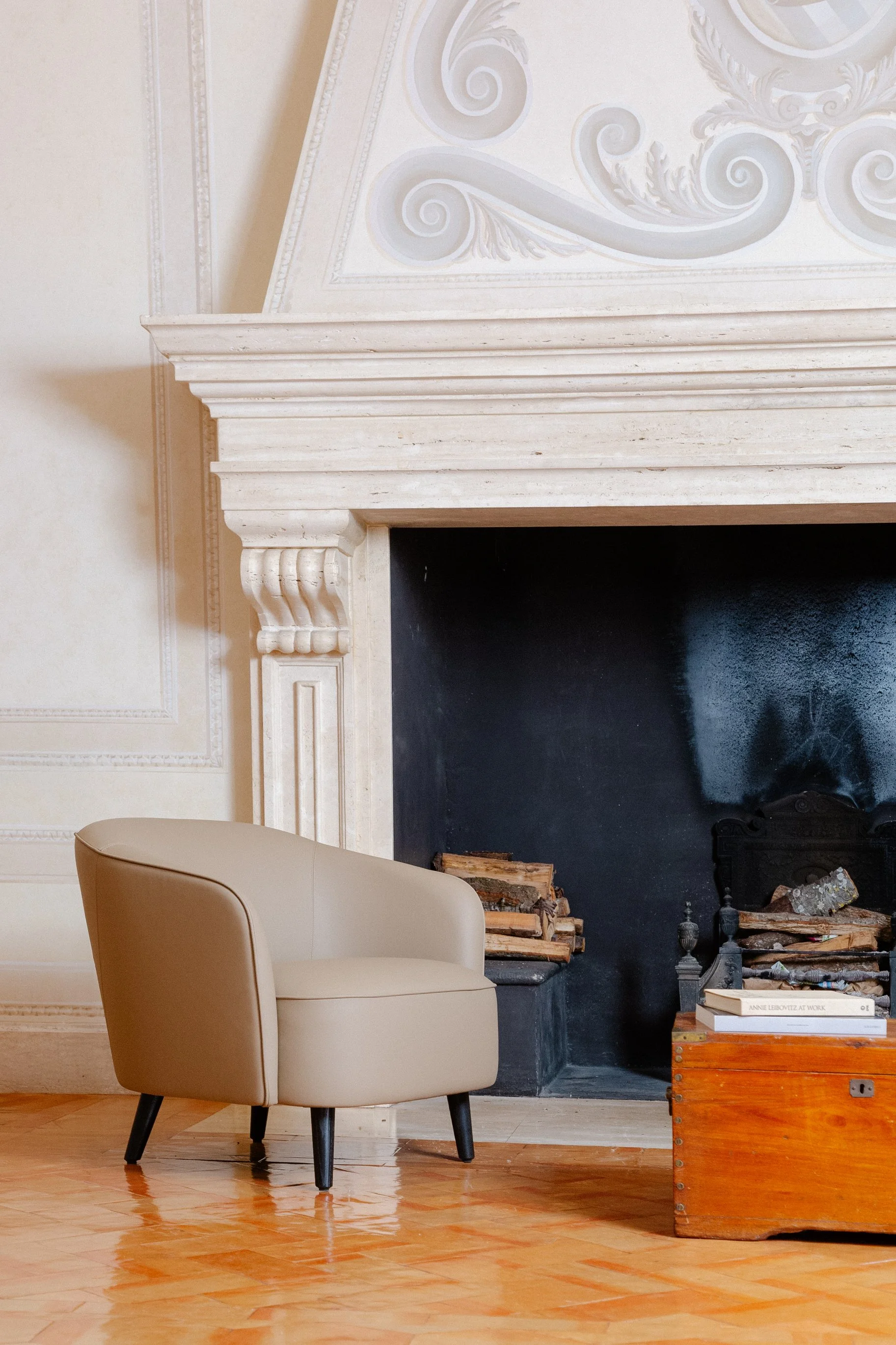 A cozy living room corner with a beige armchair, a white fireplace with ornate details, a wooden chest with books and logs, and a hardwood floor.