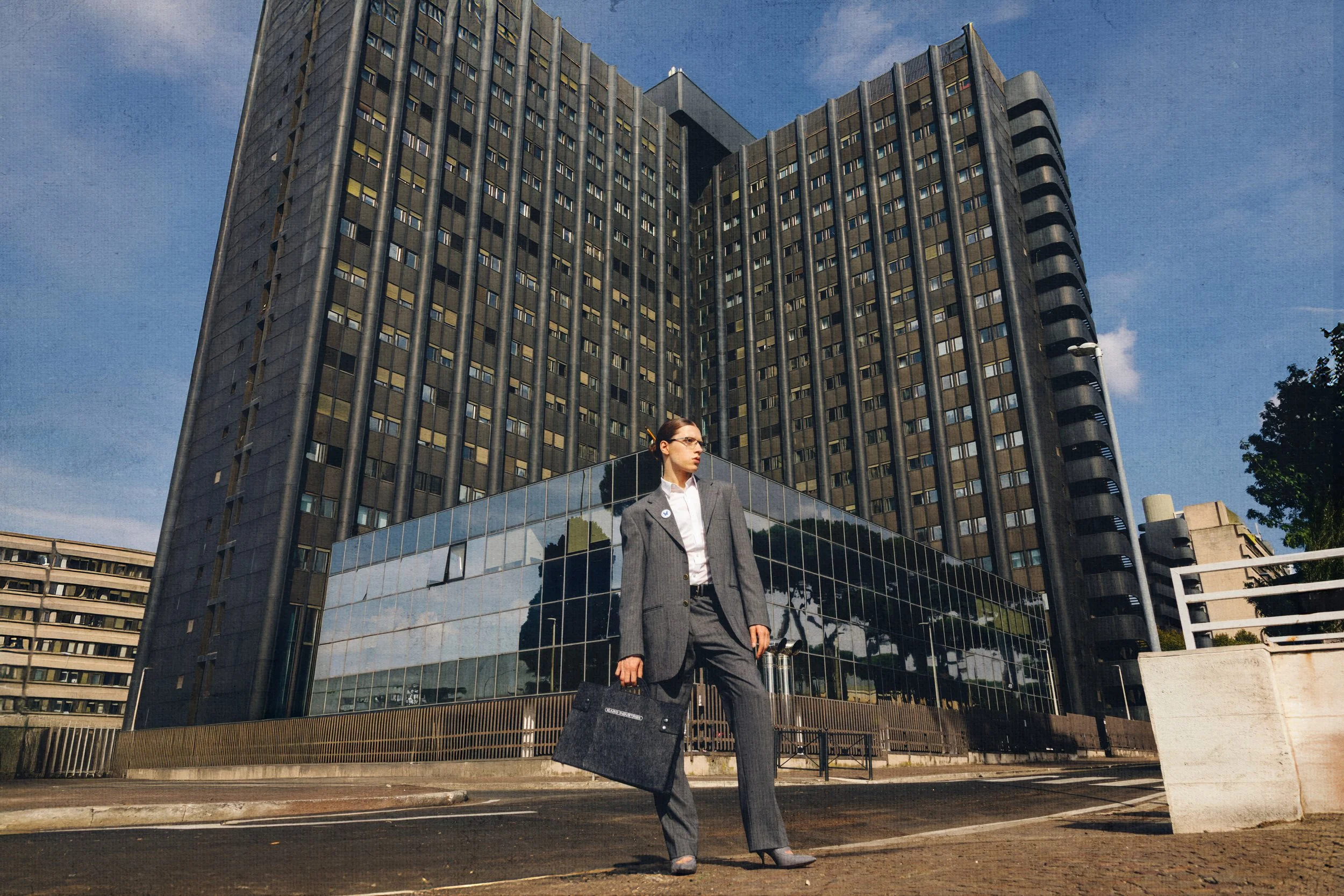 A woman in a gray business suit holding a briefcase standing on the street in front of a tall black office building with reflective glass windows.