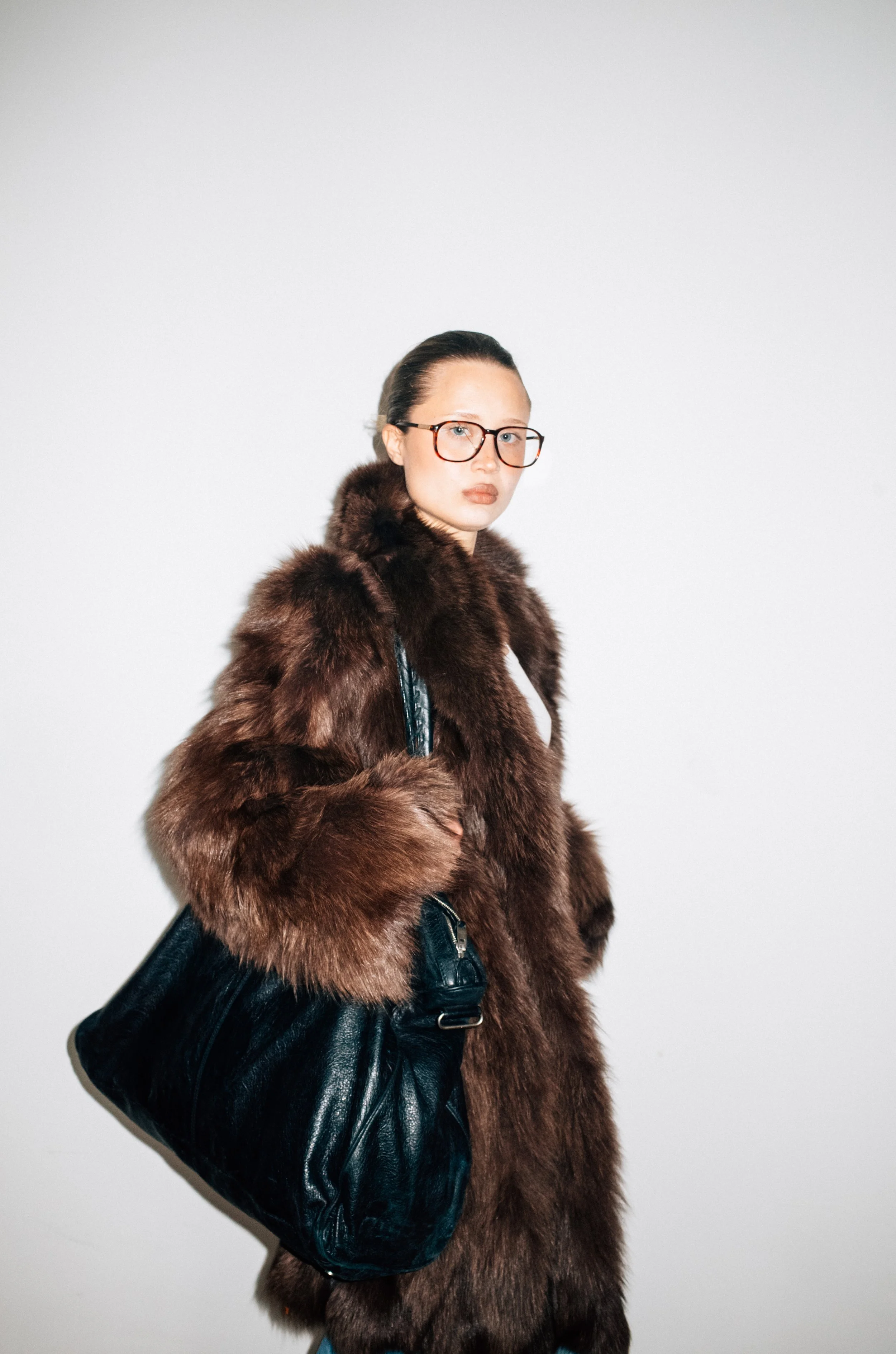 A woman wearing glasses and a luxurious brown fur coat, carrying a large black leather handbag, standing against a plain white background.