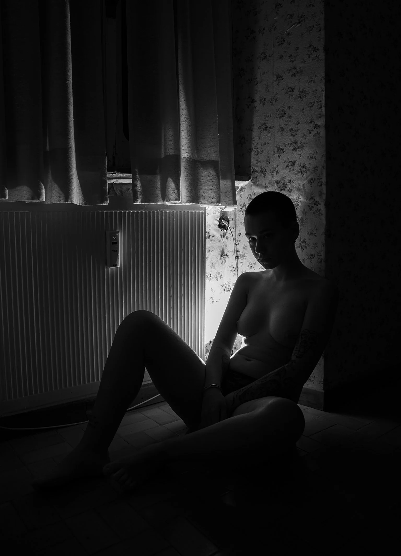 Silhouette of a topless woman sitting on the floor in front of a window with curtains, with light coming through the window, casting shadows.