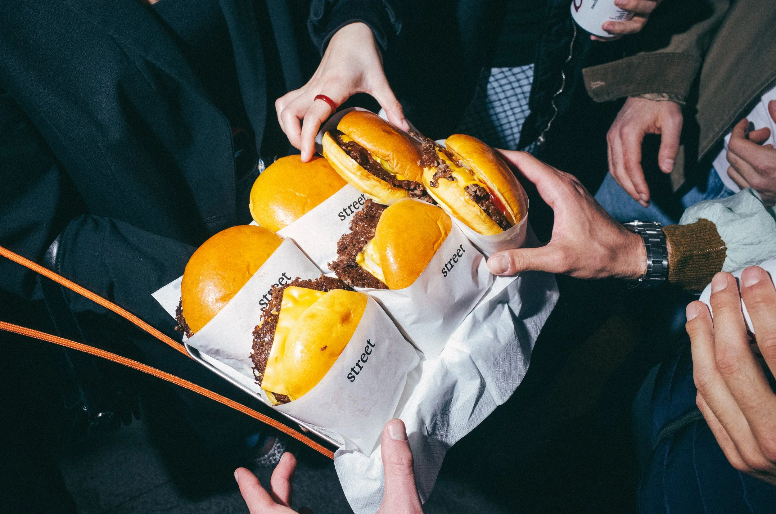 Several people sharing a tray of cheeseburgers wrapped in paper labeled 'street,' with some holding the tray.