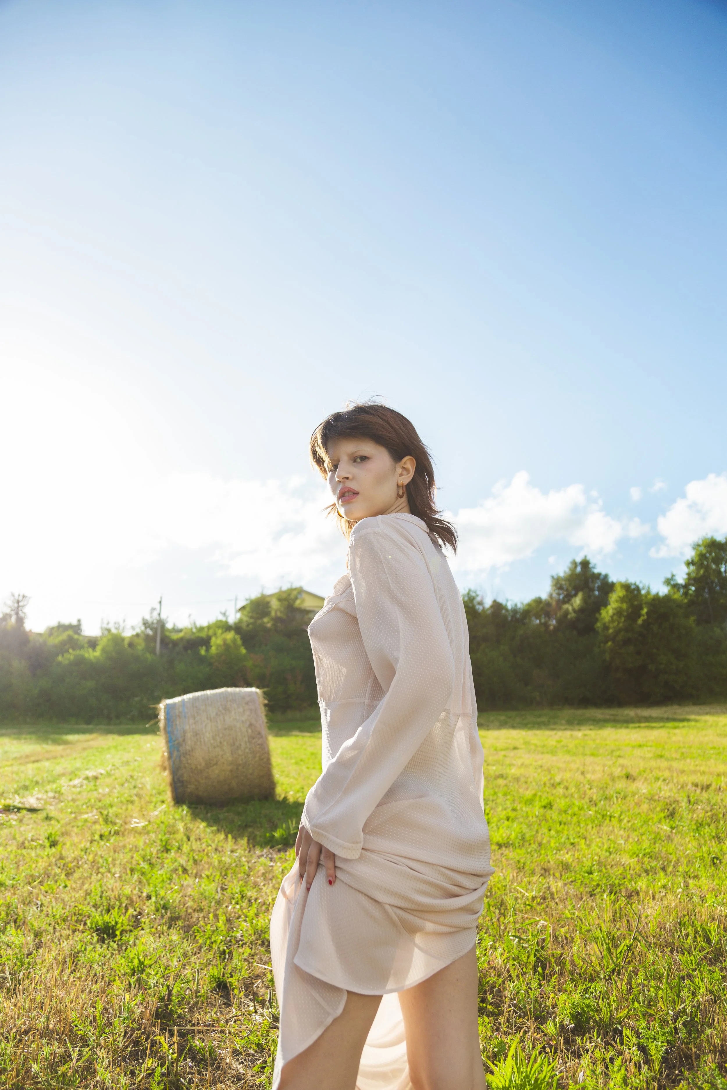 A woman standing outdoors in a field with a hay bale in the background and a bright blue sky with a few clouds.