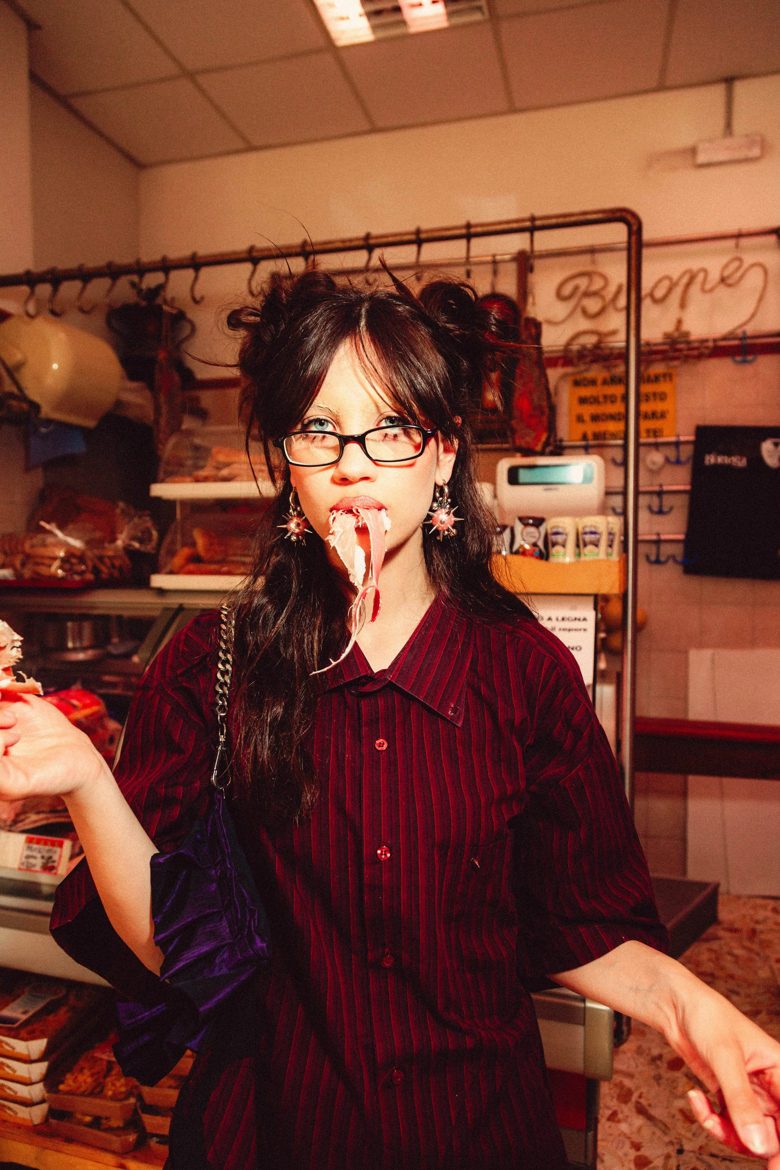 Young woman with glasses and earrings, wearing a dark red and black striped shirt, with messy hair styled in buns, is in a grocery store with meat products in the background, holding a piece of cooked meat or seafood in her mouth with a playful expre