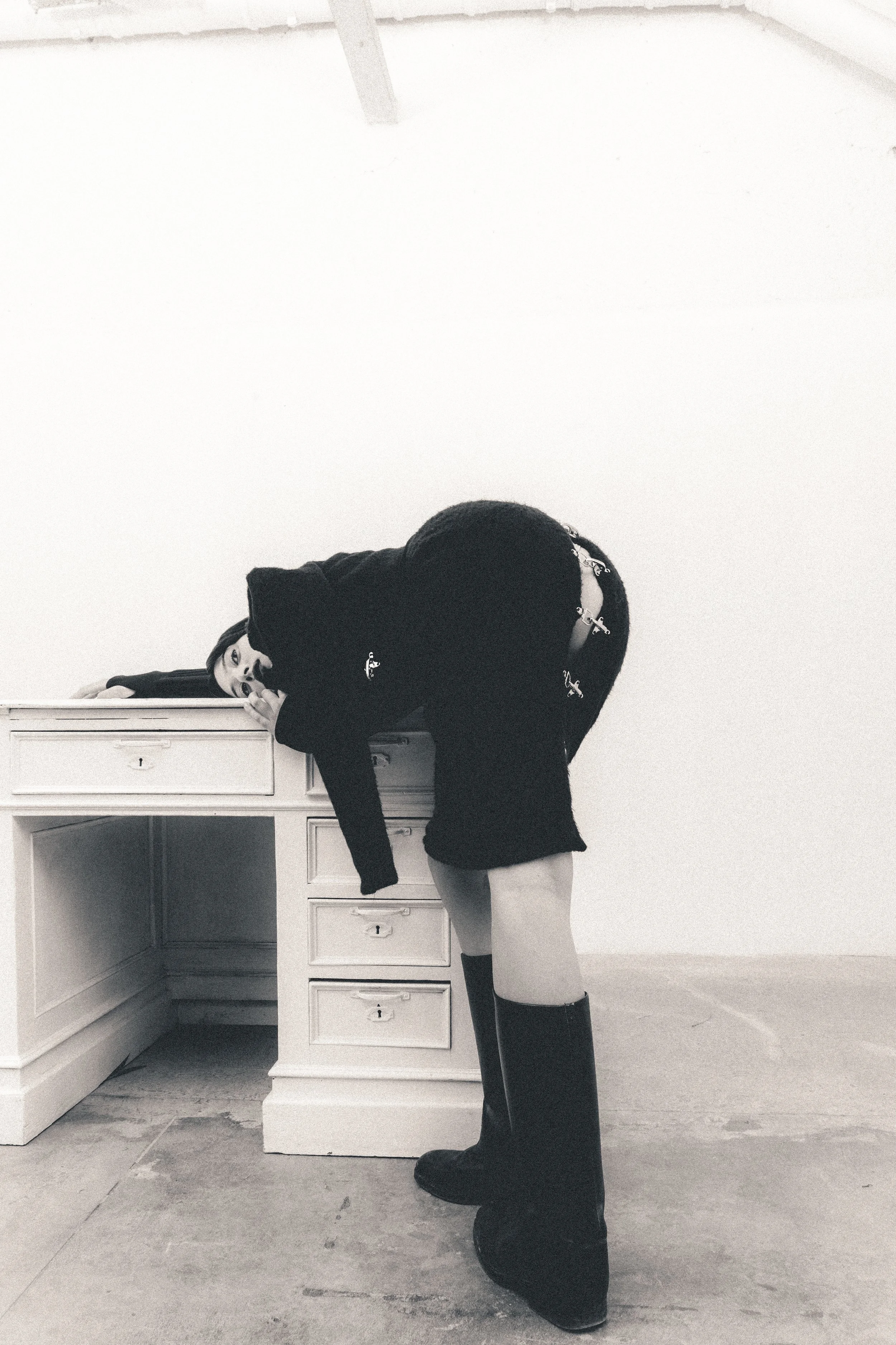A woman with long dark hair leaning on a white desk, wearing a black coat and high boots, in a sparsely decorated white room.