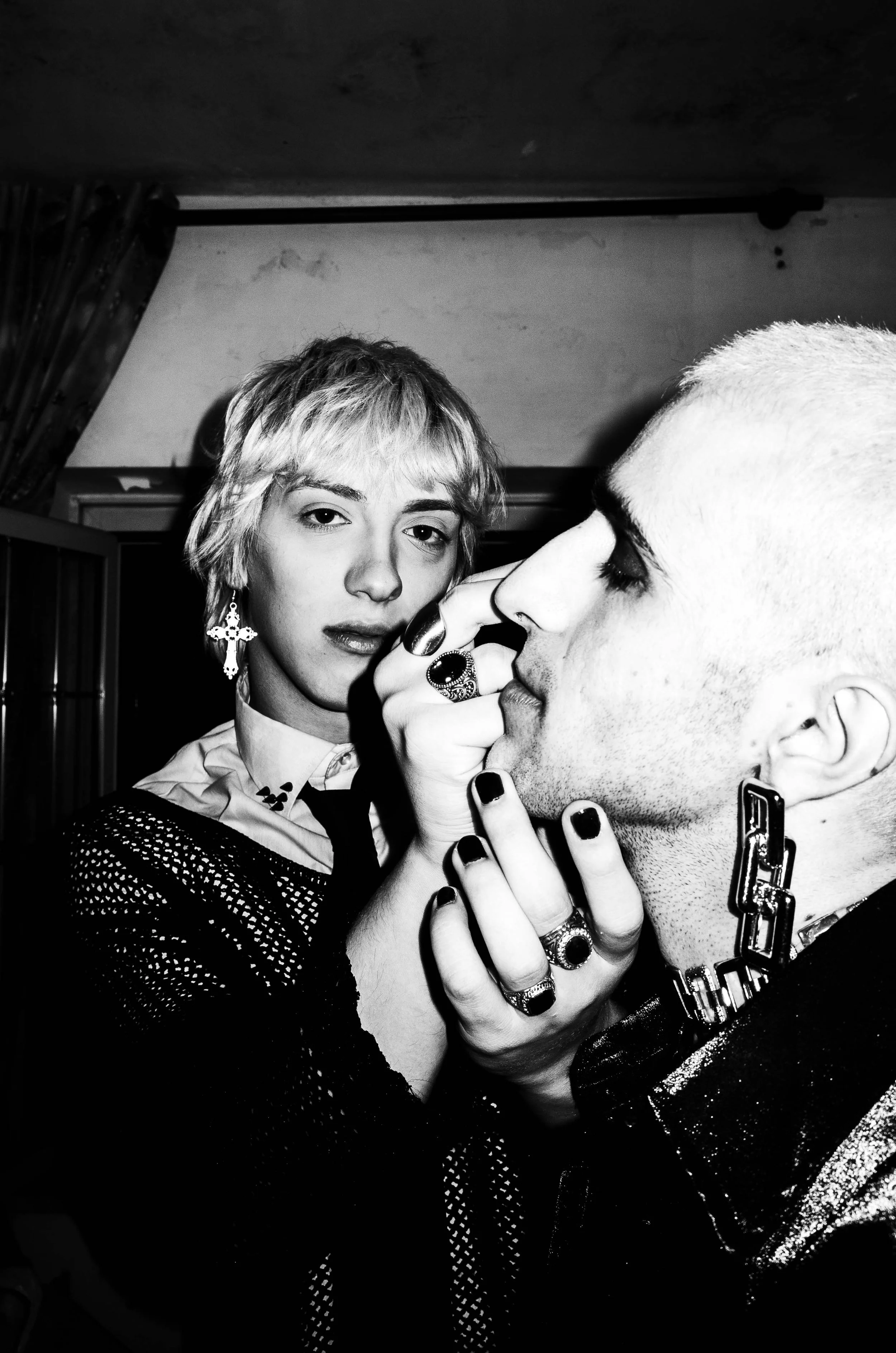 A black and white photo of a woman with short hair and earrings touching the nose of a bald man with jewelry, both with intense expressions.