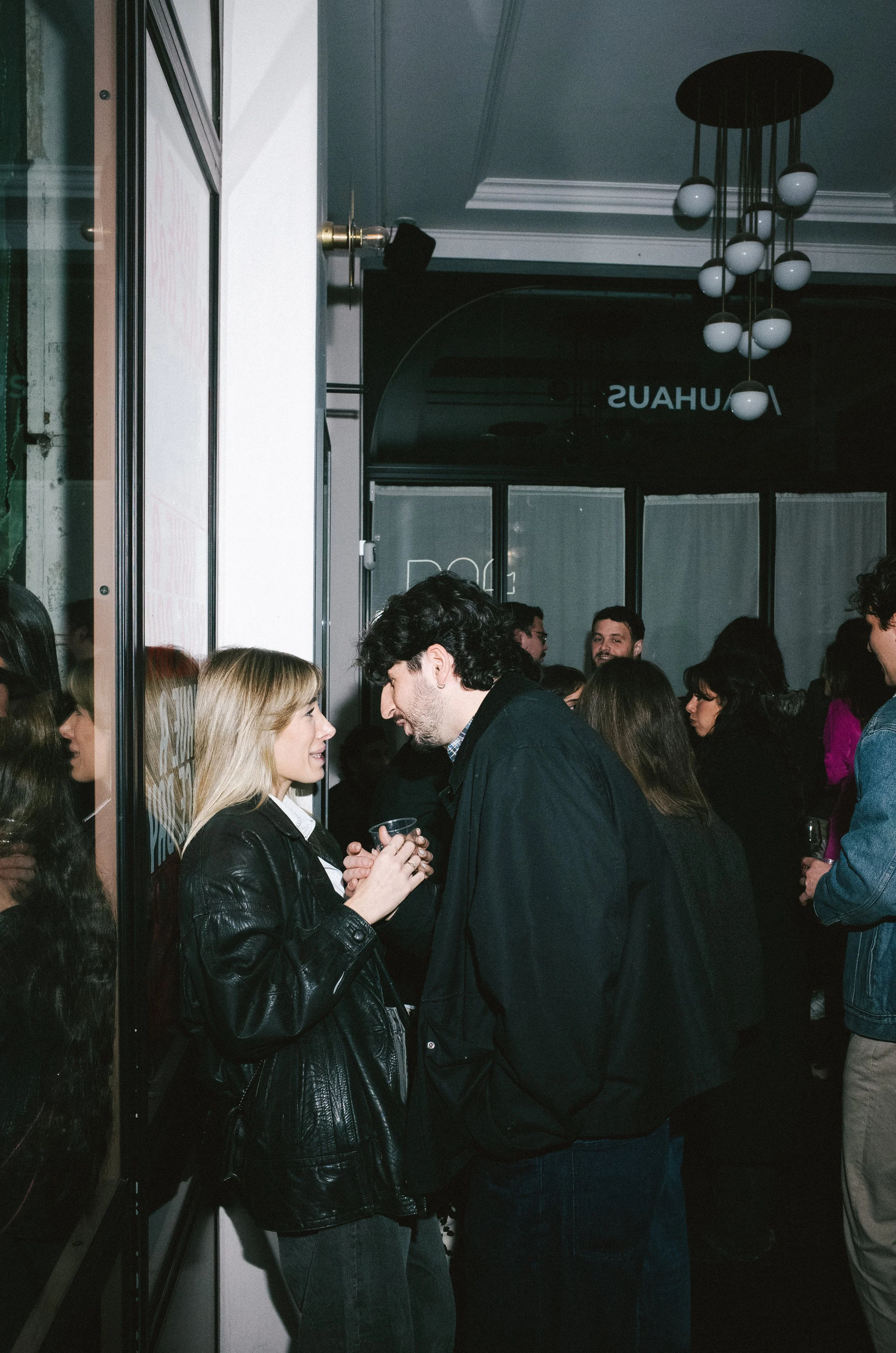 Two people having a conversation at a social event, surrounded by a crowd inside a building.