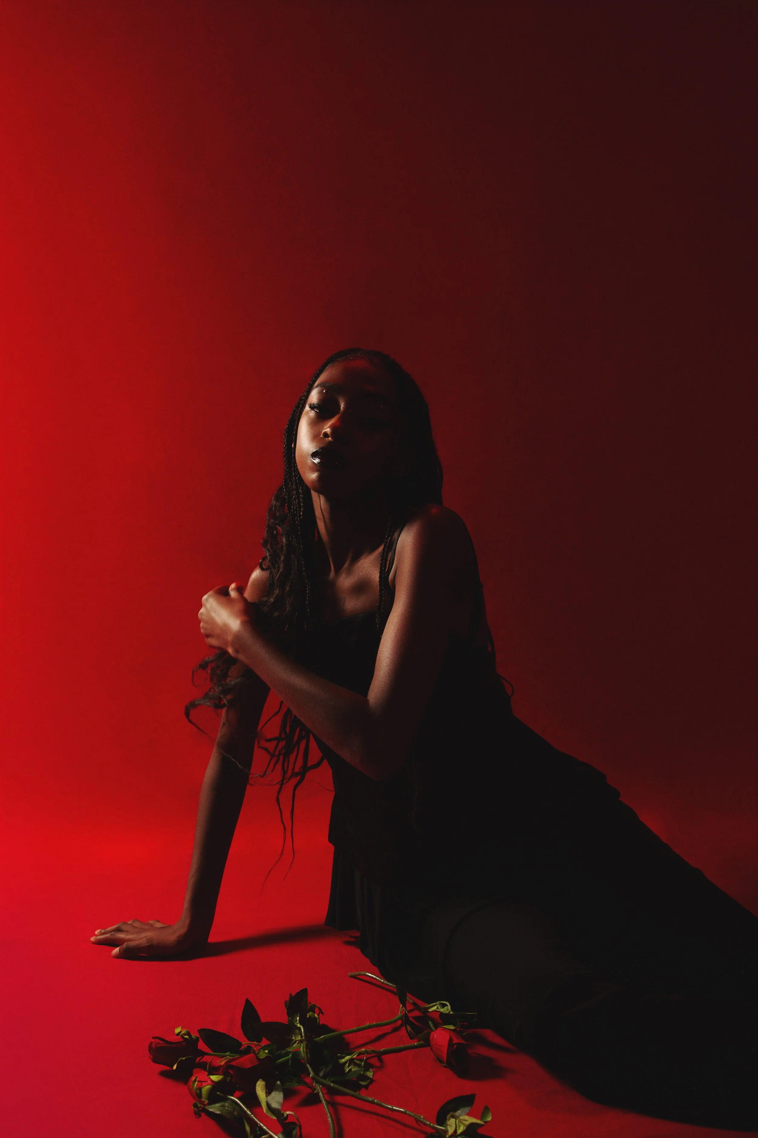 A woman with long braided hair in a black dress sitting on a red surface, with a dark red background and a bouquet of roses in front of her.