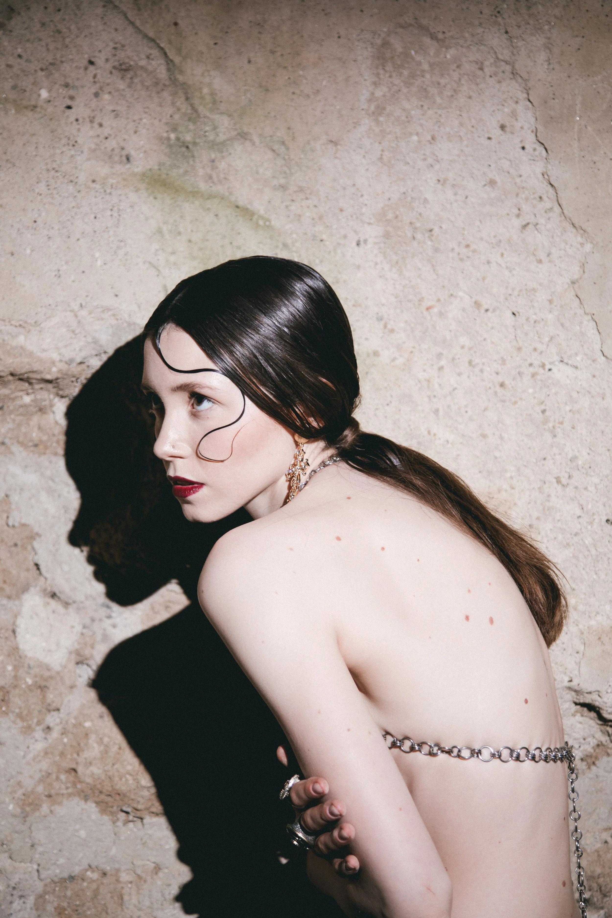 A woman with dark hair in a ponytail, wearing earrings and jewelry, poses against a rough, textured wall with a shadow cast on the wall behind her.