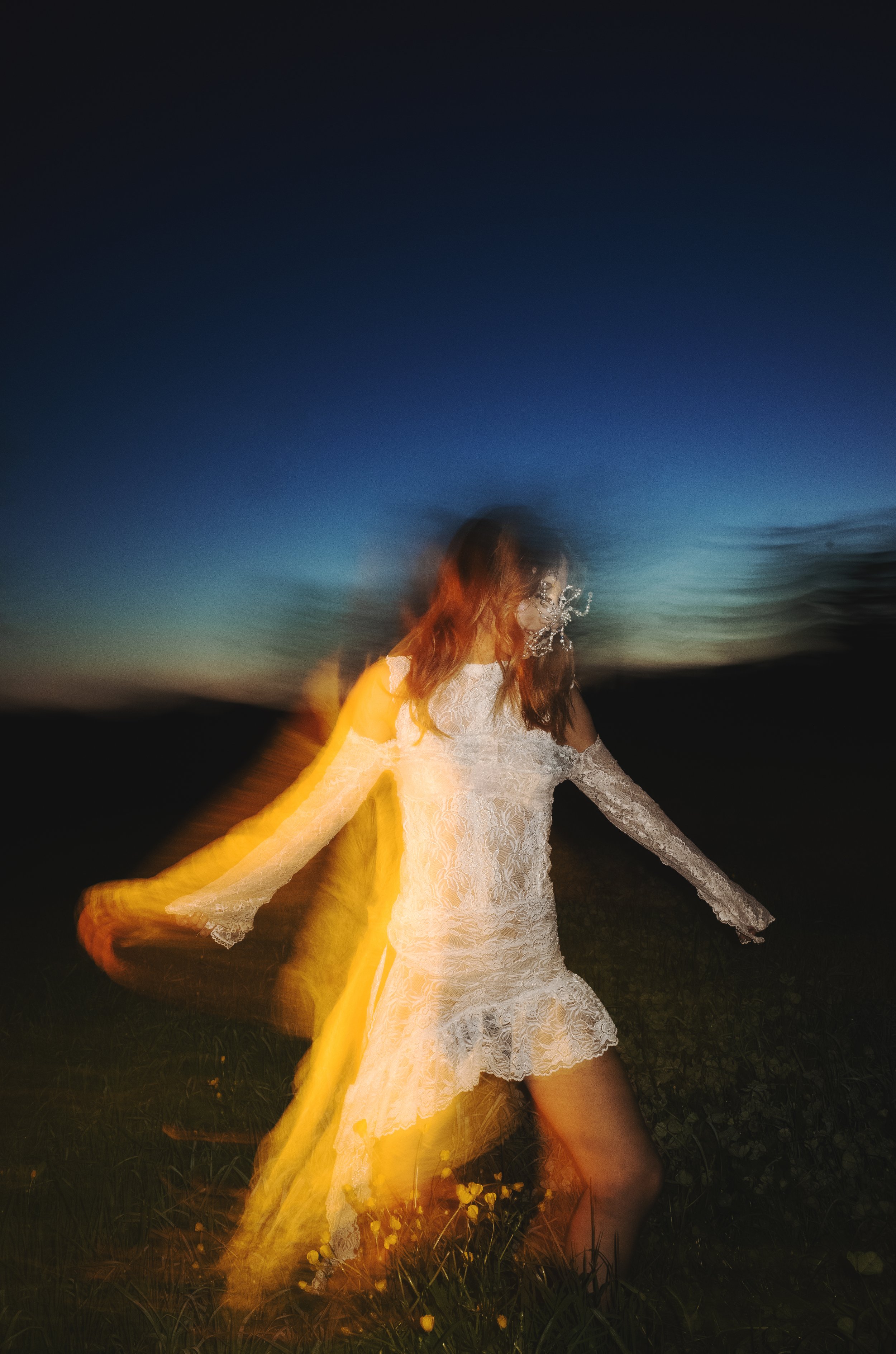 A woman in a white lace dress stands outdoors at dusk, with motion blur and light trails around her, creating a surreal effect.