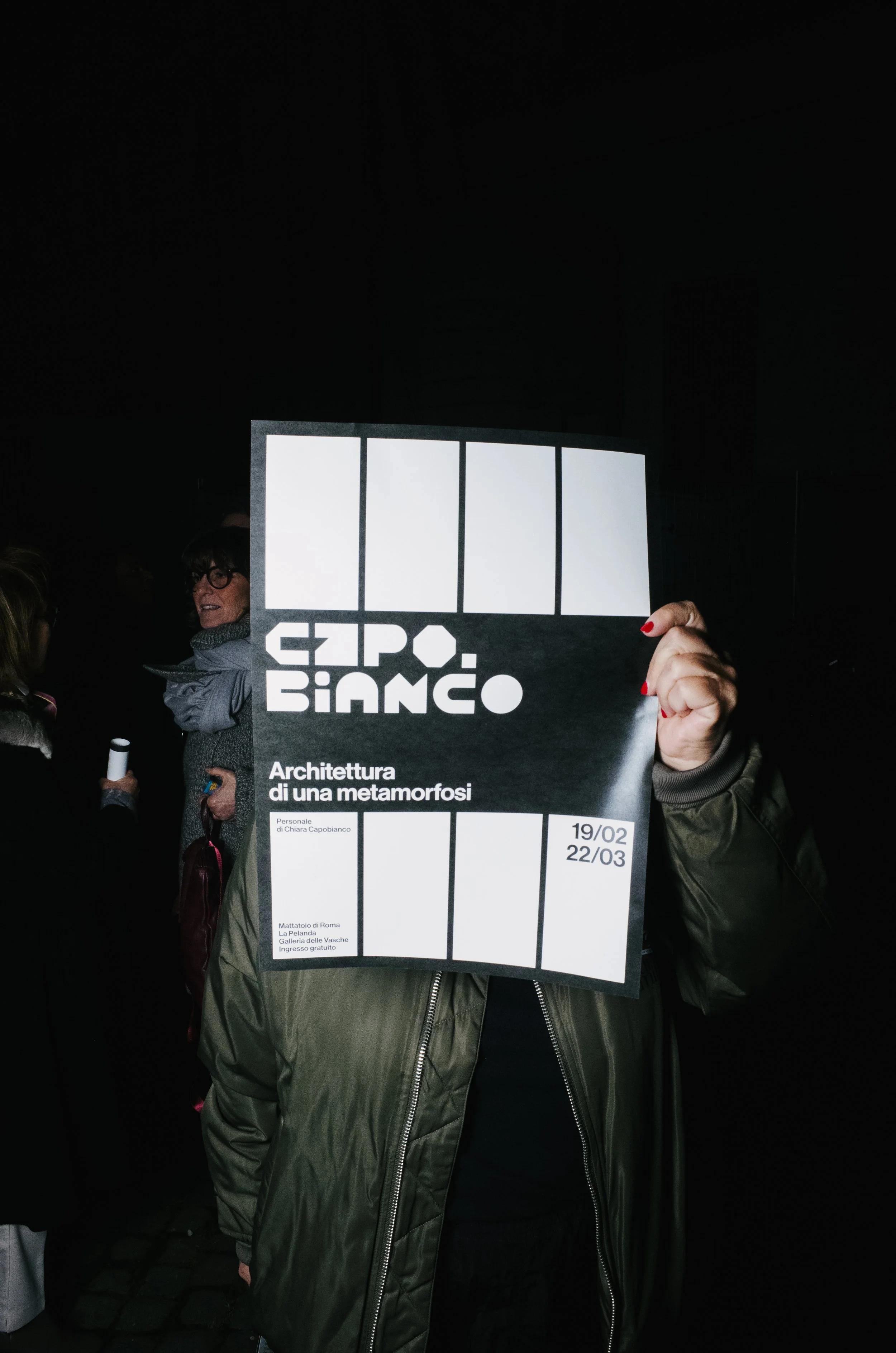 Person holding a poster for the Expo Bianco event, titled 'Architettura di una metamorfosi,' with dates February 19 to March 22, and details of the venue in Rome, Italy.