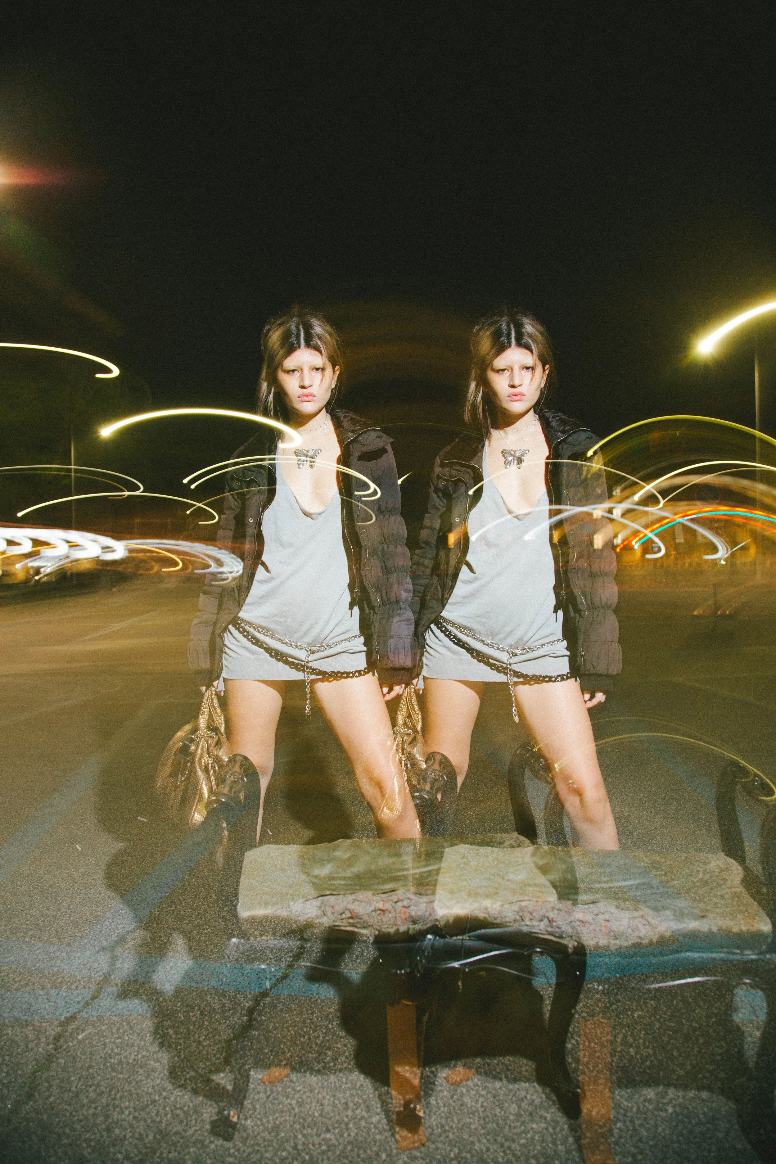 Nighttime photo of two young women standing on a street with light streaks and reflections, wearing casual streetwear with jackets, skirts, and handbags.