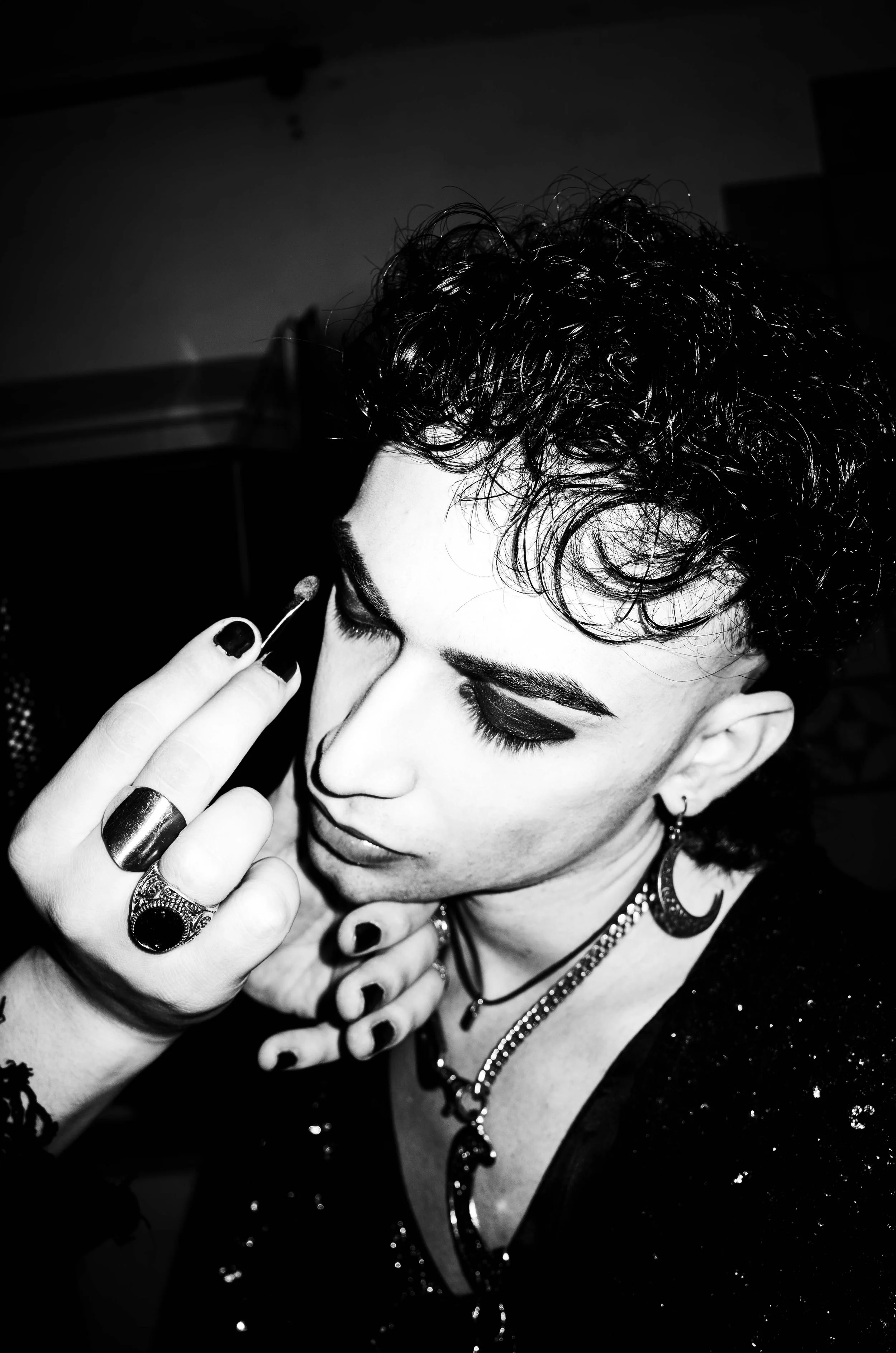 Person applying makeup with a cotton swab, wearing dark eye makeup, jewelry, and a sparkly top, in black and white.