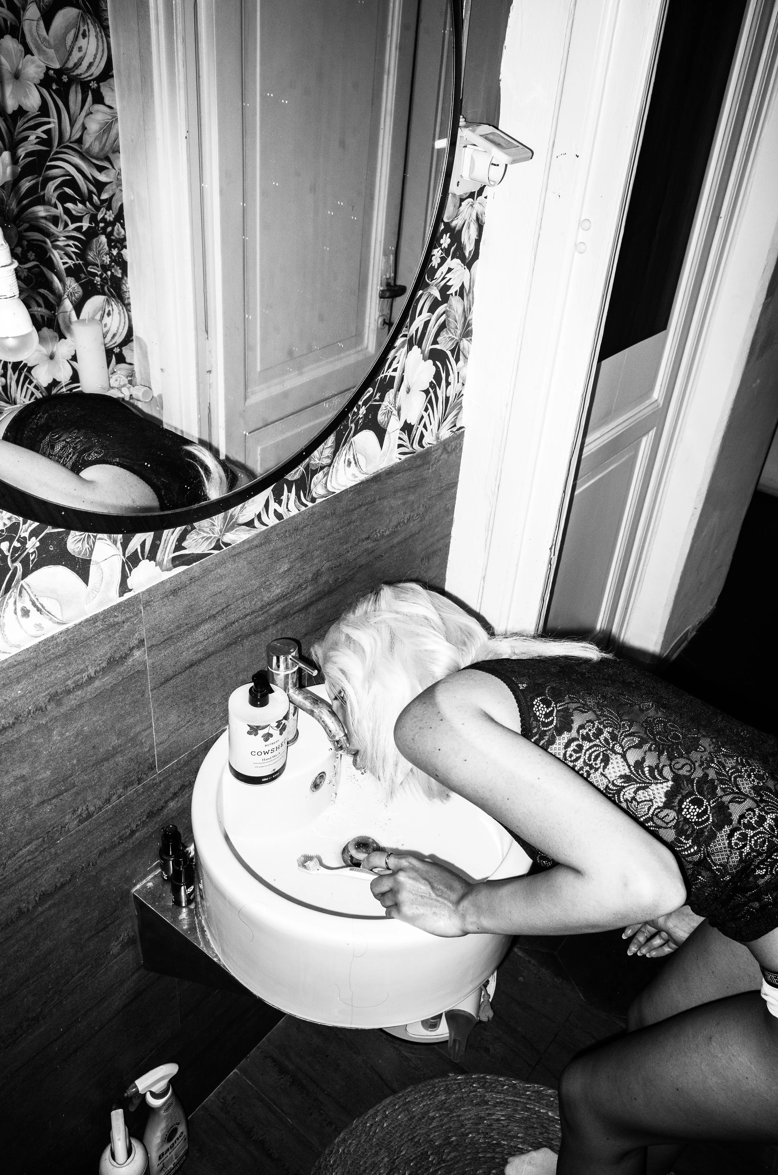 A woman with lace top leaning over a small sink, her head at the sink's faucet, with a dog drinking from the sink.