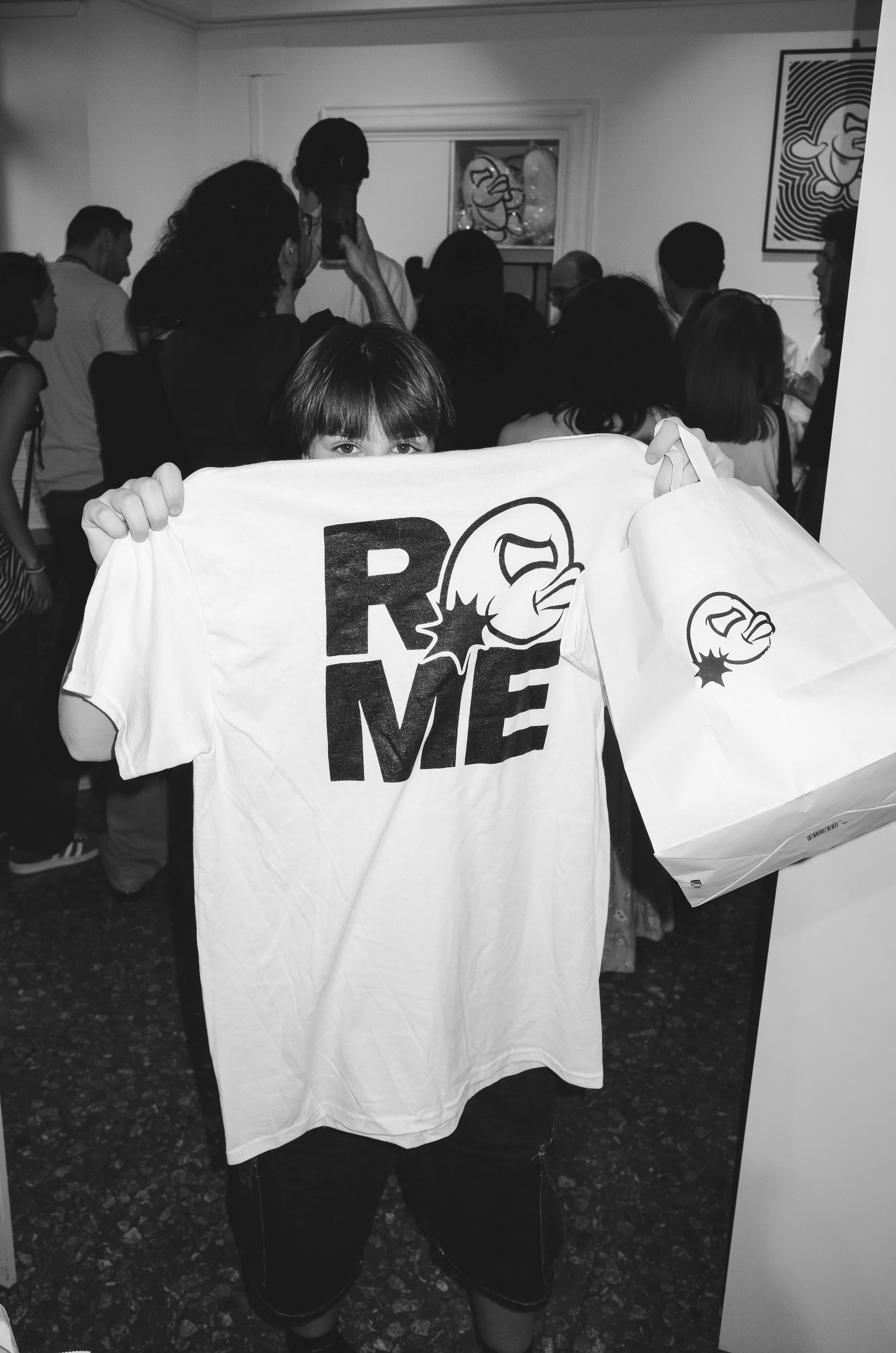 A young girl with short hair holding a white t-shirt and a shopping bag with a comic-style angry face and the word "RAGE" printed on them, in a crowded room with art on the walls.