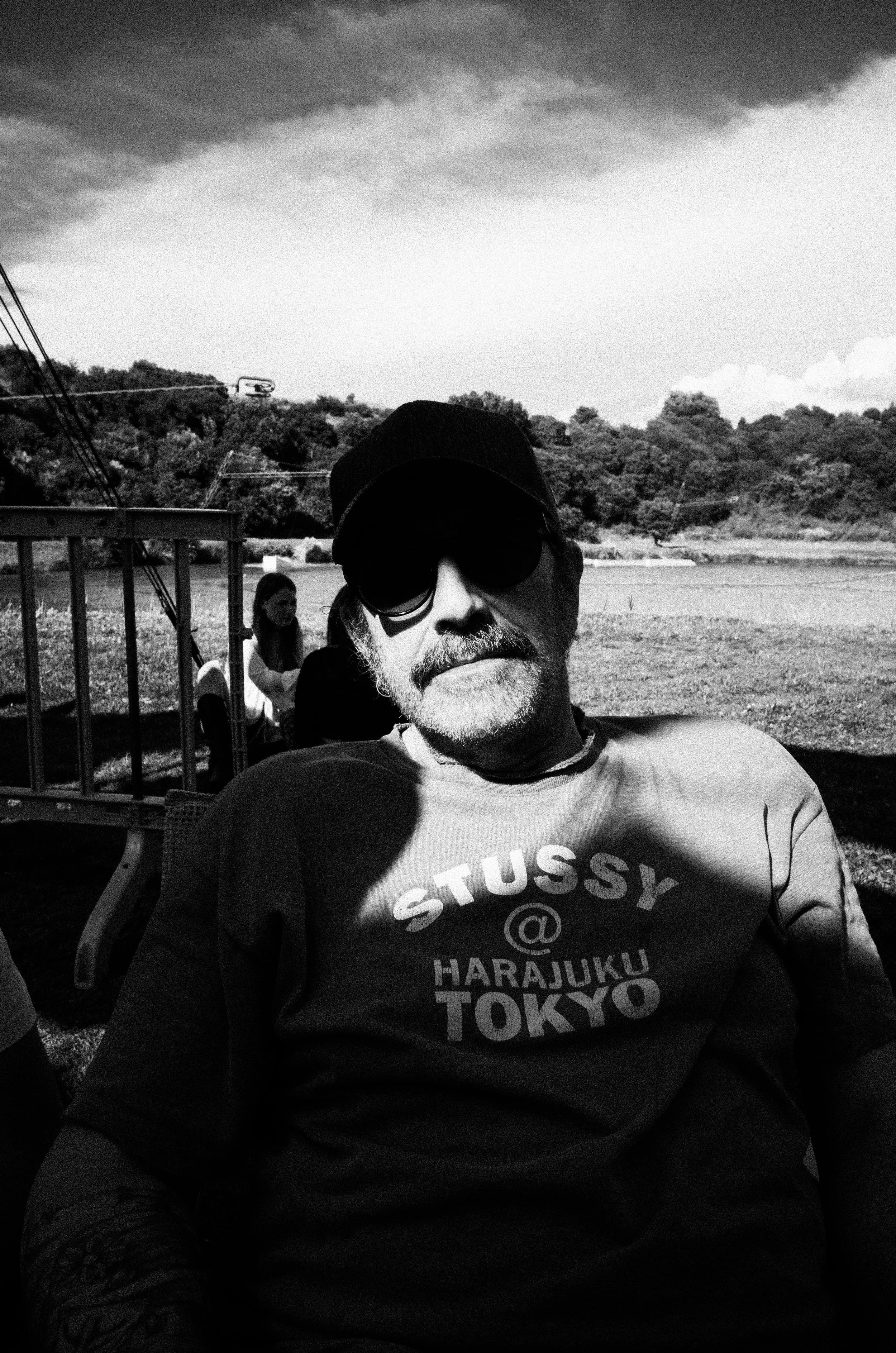 Black and white photo of a man wearing sunglasses, a cap, and a T-shirt with the text 'Stussy @ Harajuku Tokyo' sitting outdoors with a scenic landscape and trees in the background. There is a woman in the background sitting near a wooden structure.