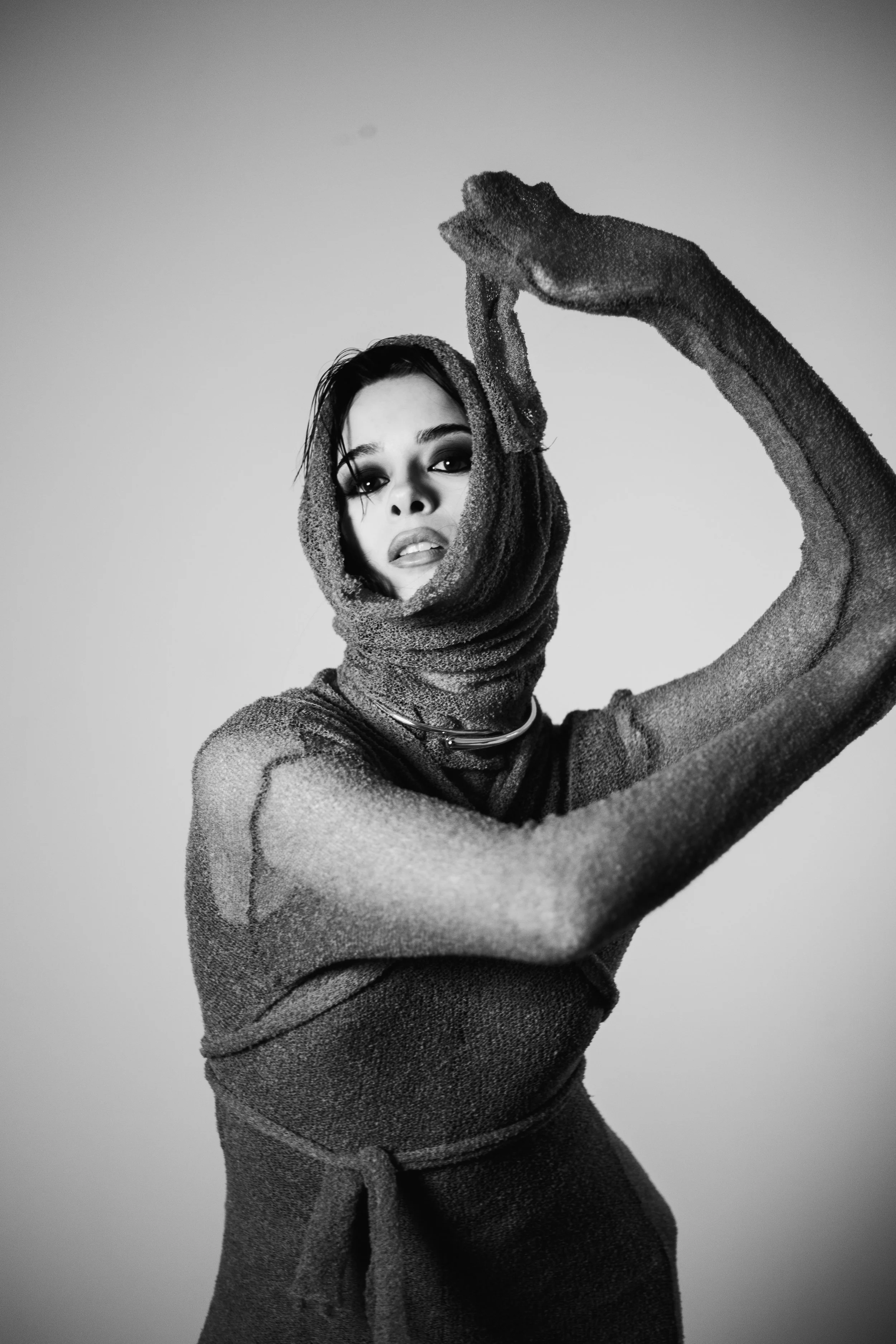 Black and white portrait of a woman wearing a textured fabric wrap around her head and body, with dark eye makeup, posing with one arm raised and bent, holding her head with her hand.