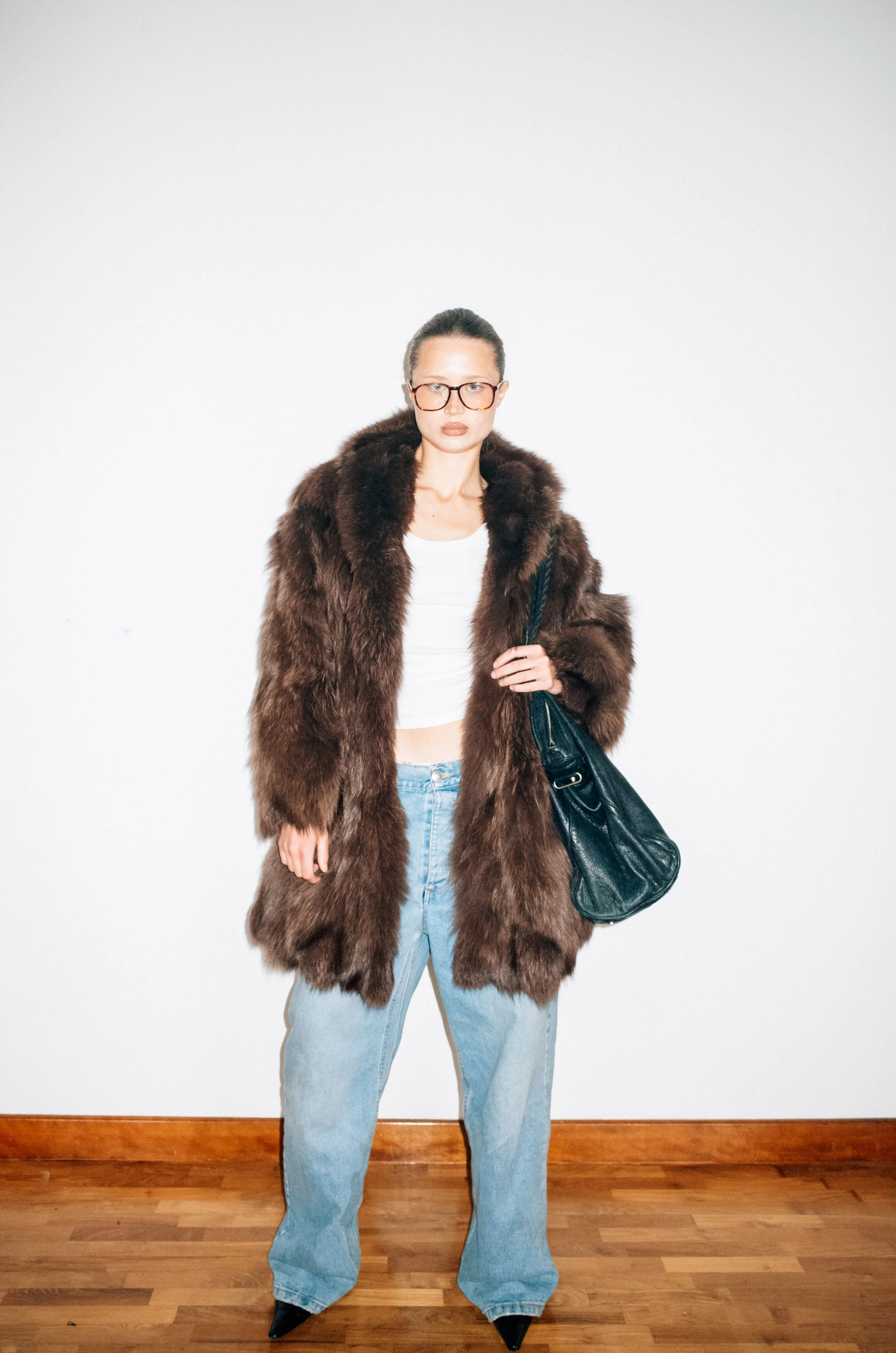 Woman wearing glasses, a large brown fur coat, a white crop top, loose blue jeans, black pointed shoes, and carrying a black handbag, standing against a plain white wall and hardwood floor.