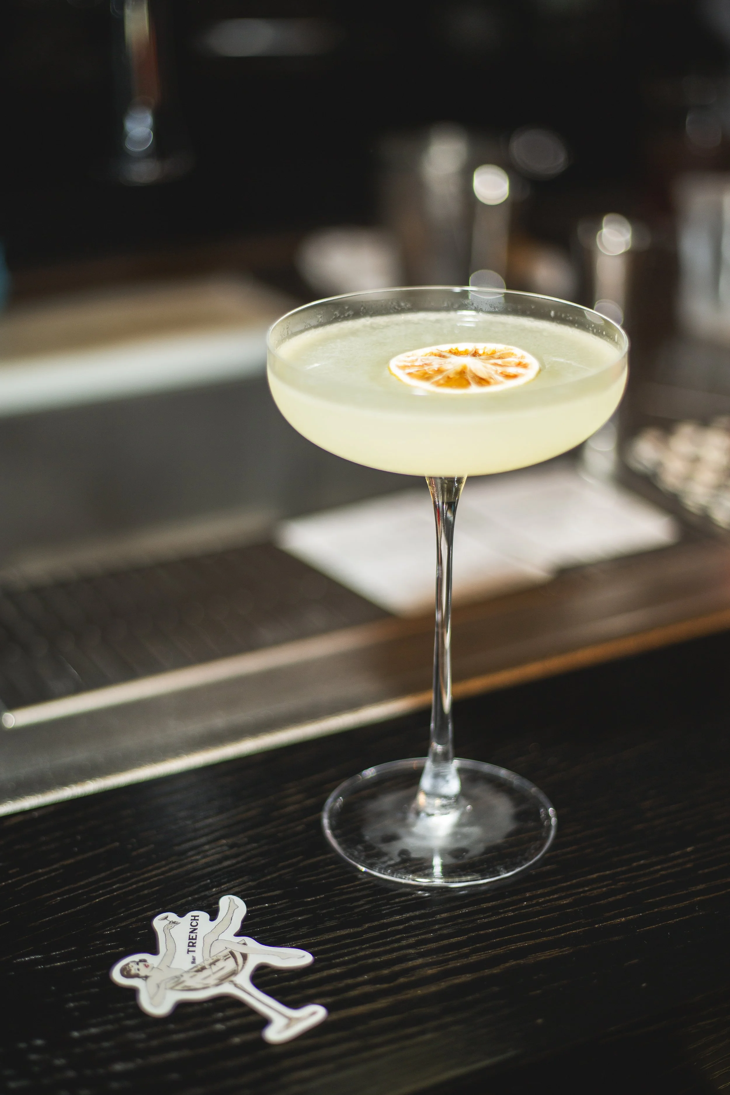 A cocktail glass with a yellow-green drink, garnished with a slice of citrus fruit, placed on a dark wooden bar counter.