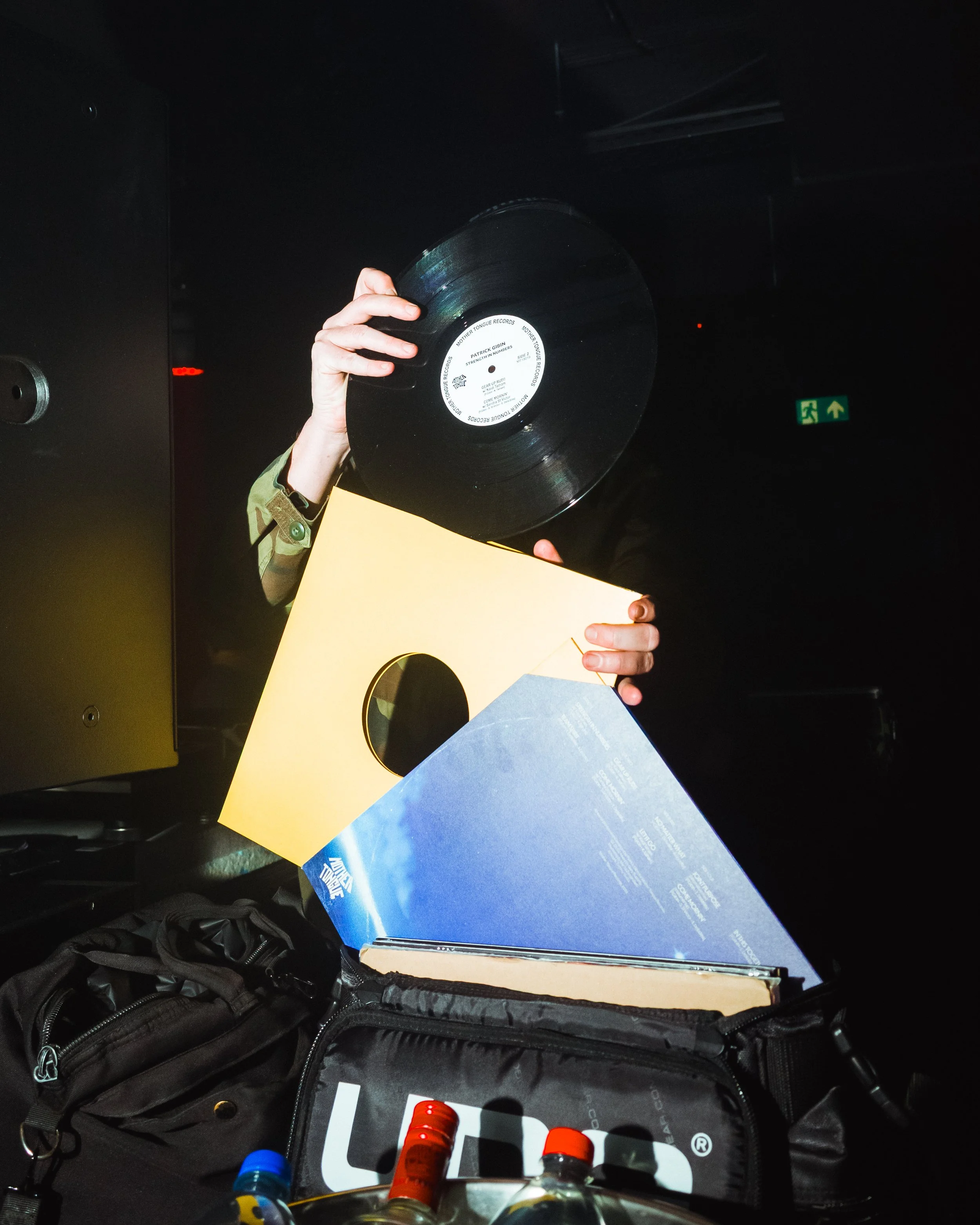 Person holding a vinyl record and a record cover with a blue sky and clouds, near a black bag with bottles inside, in a dark room with an emergency exit sign in the background.