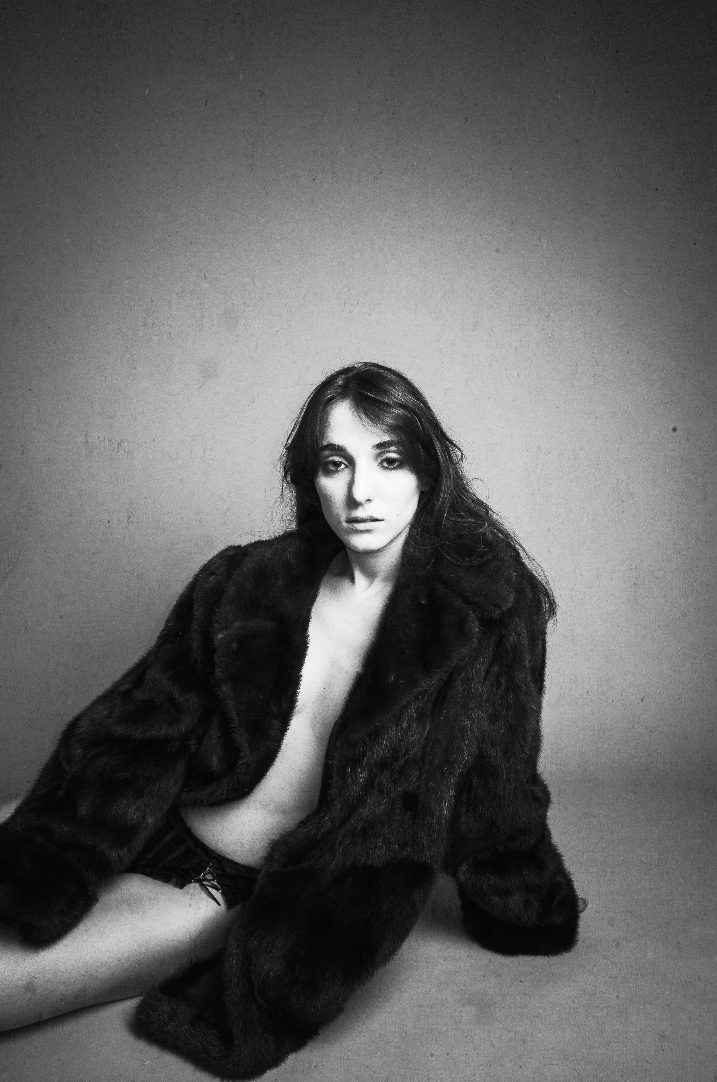 Black and white portrait of a woman with long hair, seated on the floor, wearing a fur coat and shorts