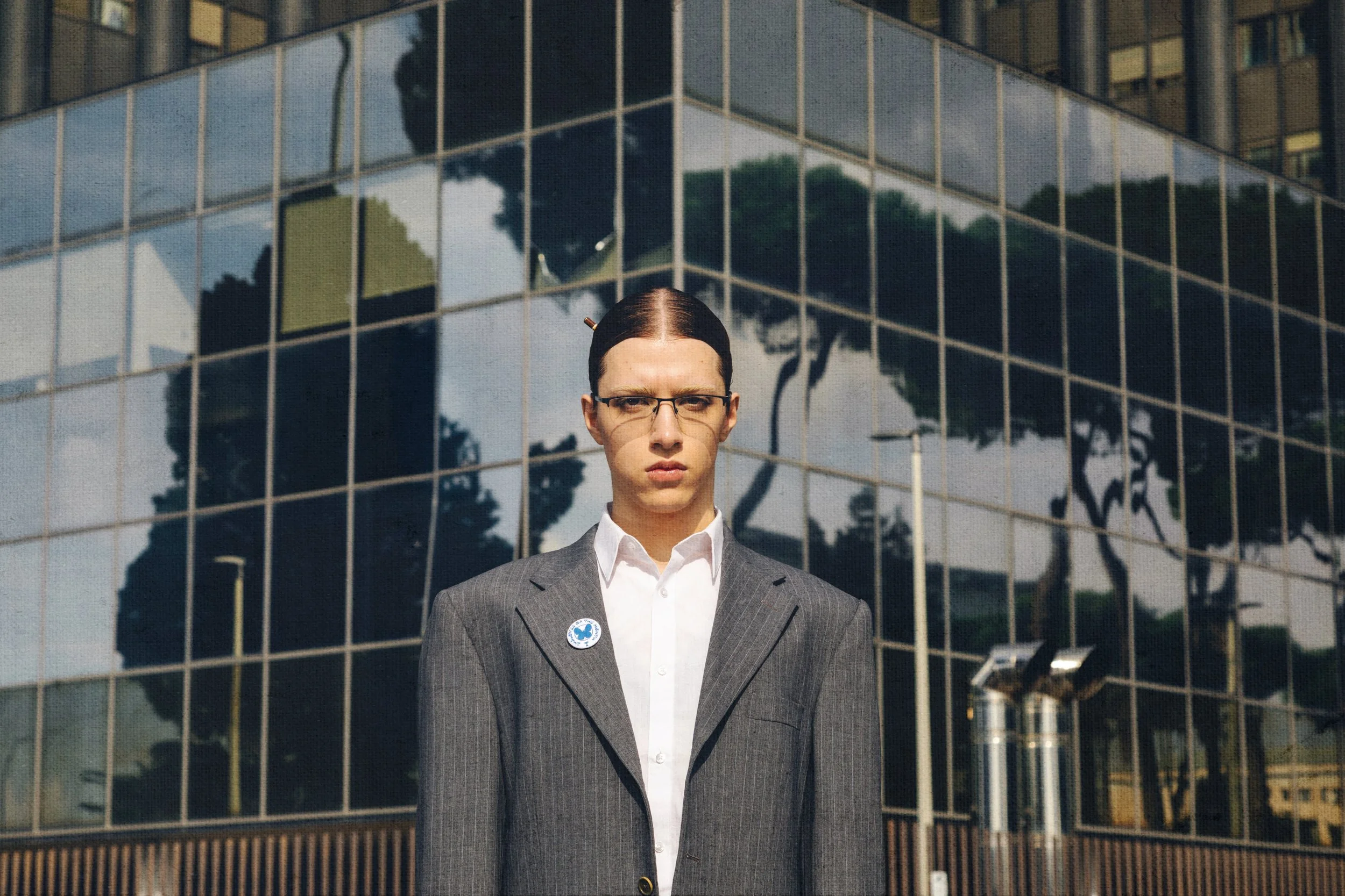 A woman with glasses and dark hair in a bun standing in front of a reflective glass building, dressed in a gray suit and white shirt, with a serious expression.