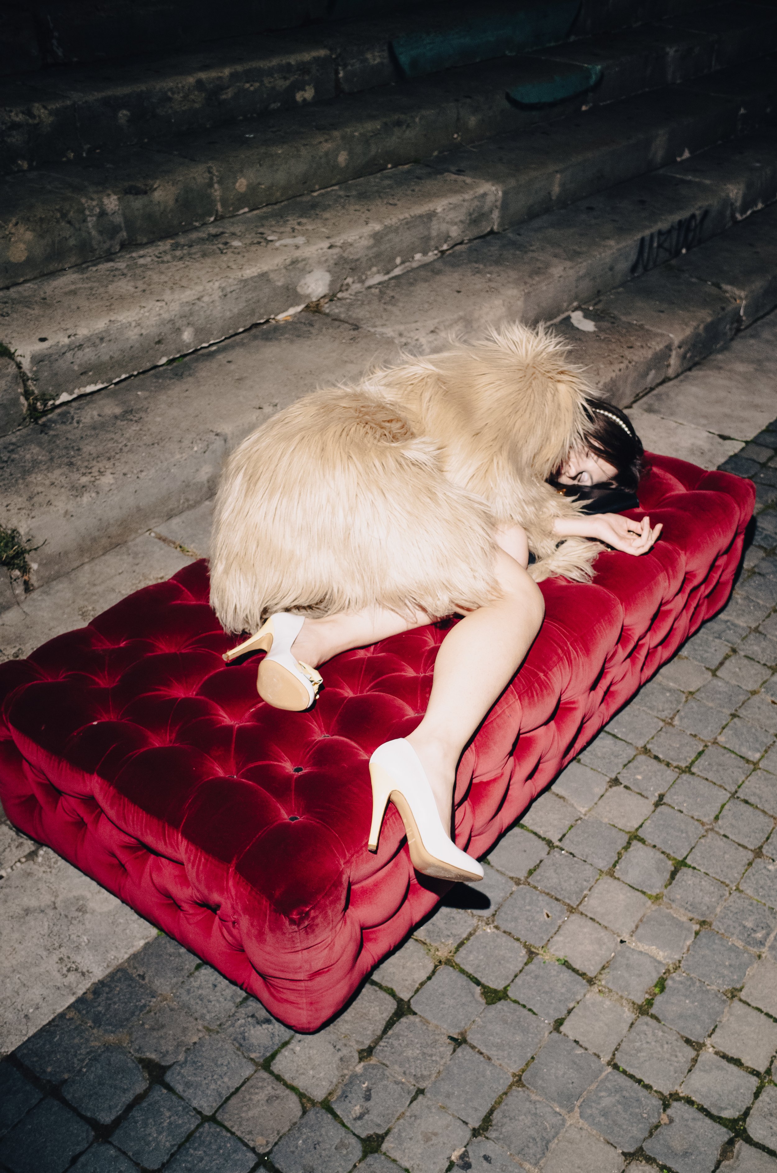 A person lying face down on a red velvet couch outdoors at night, wearing high heels and a black face mask, with their head turned to the side and a large, fluffy dog resting on top of them.