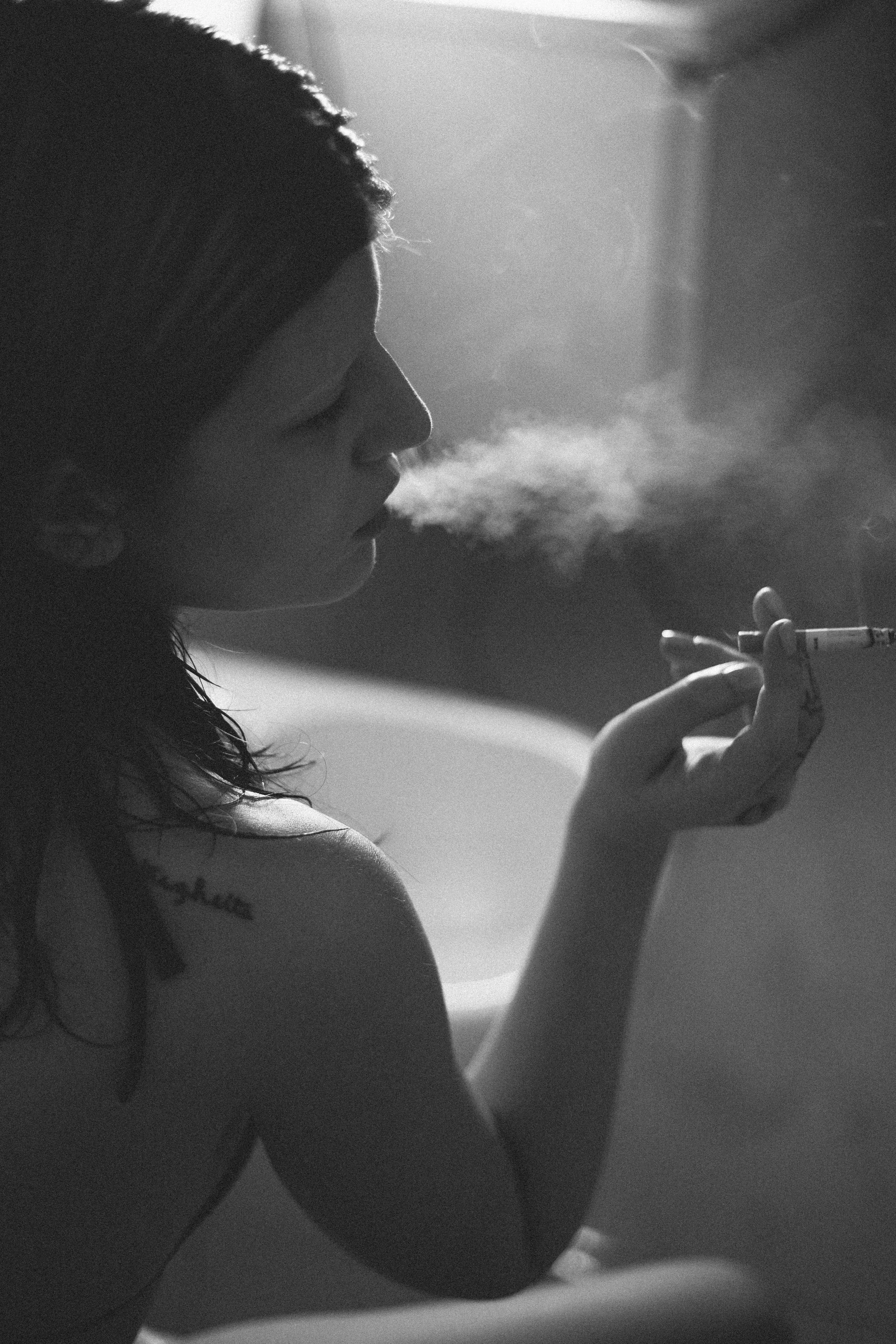 A young woman with wavy hair and tattoos on her arm is sitting and smoking a cigarette in a dimly lit room, with smoke visible in the air.