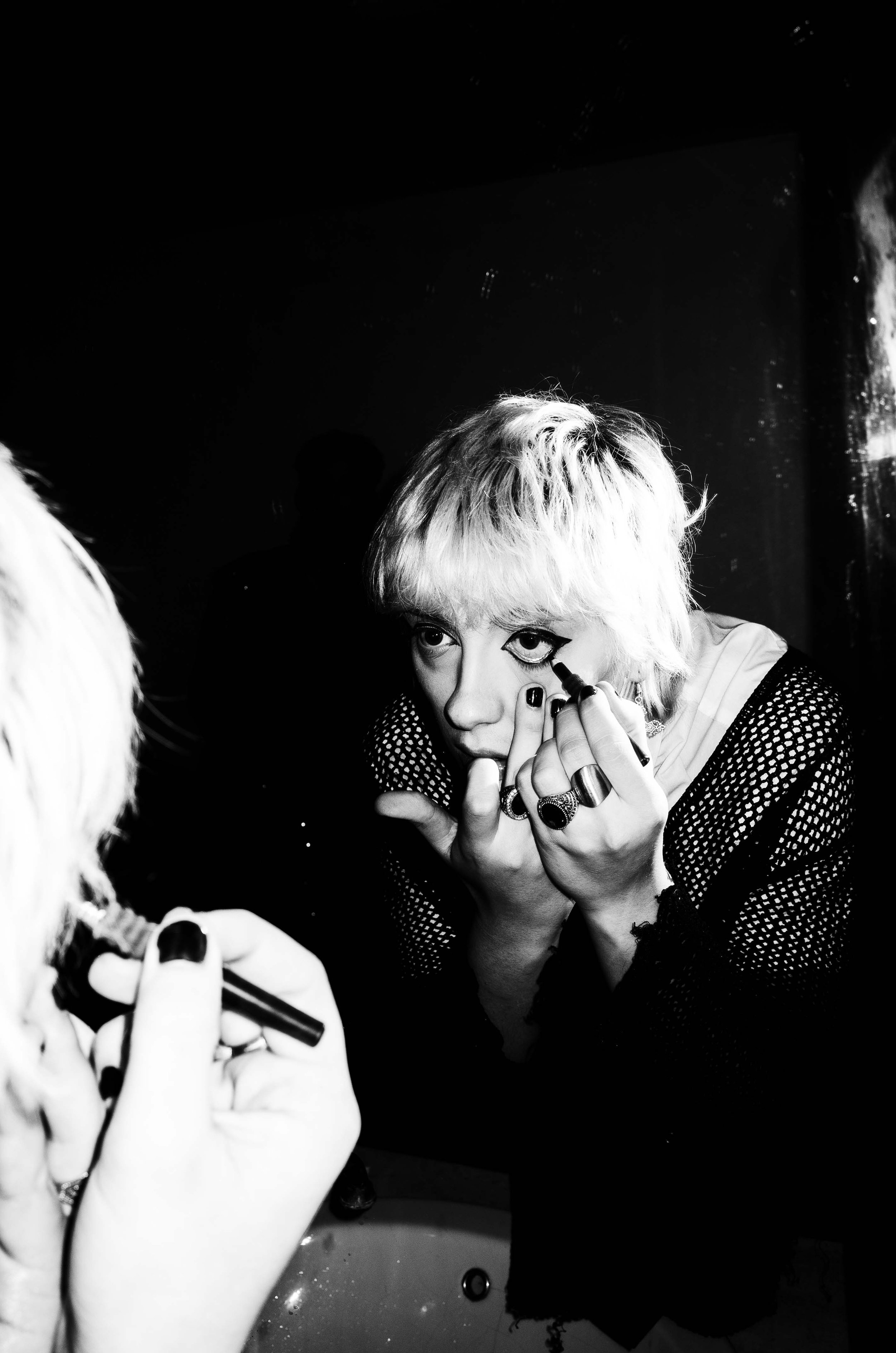 Person with light-colored hair applying black eyeliner to their lower eye in front of a mirror, wearing rings and a mesh top.
