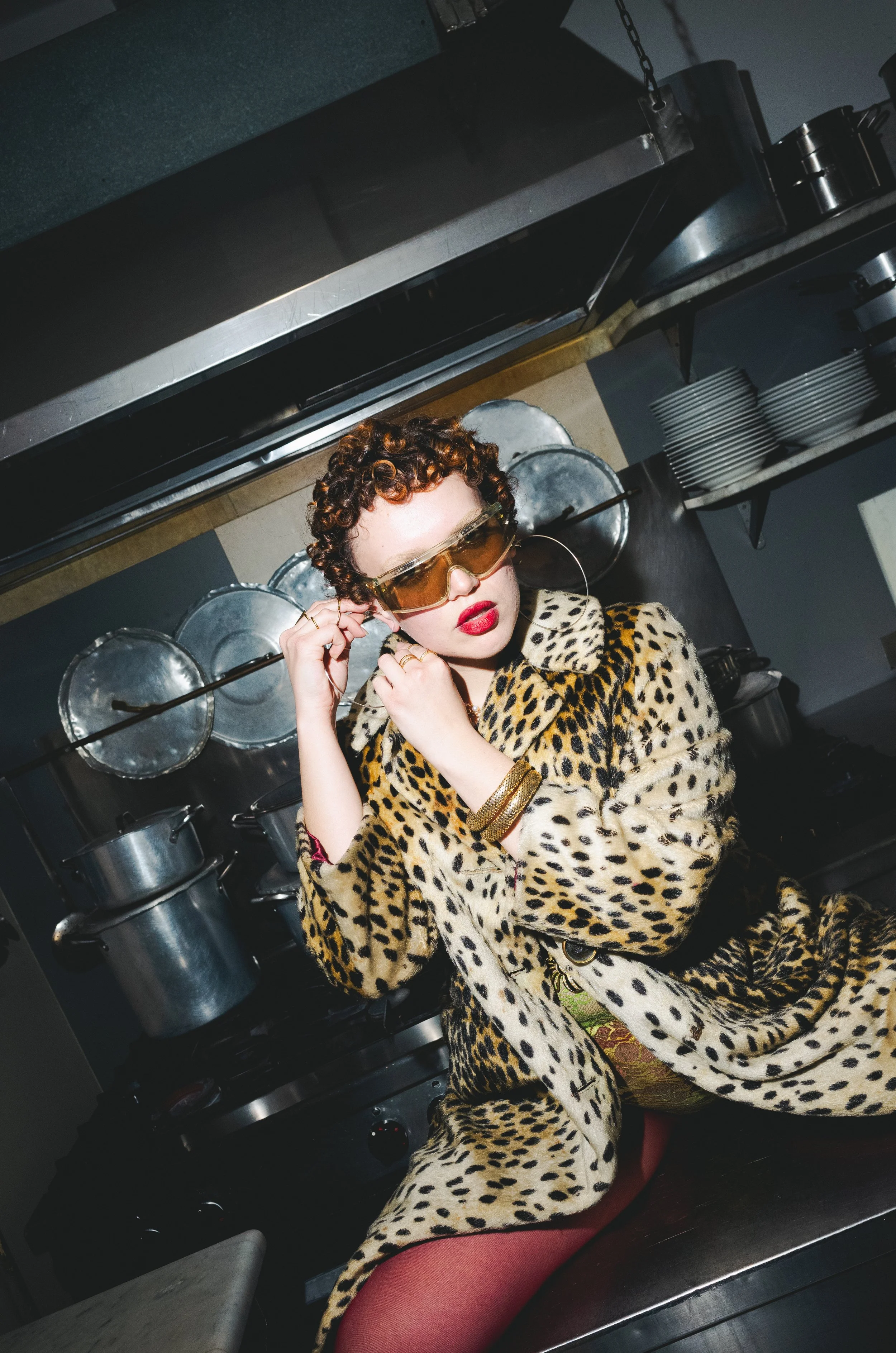 Woman with short curly hair wearing sunglasses, a leopard print coat, red lipstick, and gold jewelry in a kitchen with stainless steel pots and pans.