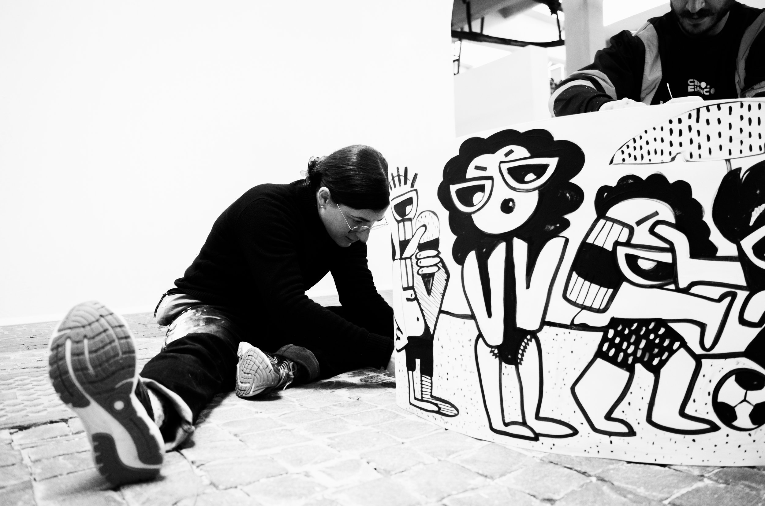 A person with glasses and long hair, dressed in dark clothing, is sitting on the brick floor and drawing on a large, colorful, cartoon-style poster featuring happy, playful characters with sunglasses, a striped shirt, and a soccer ball.