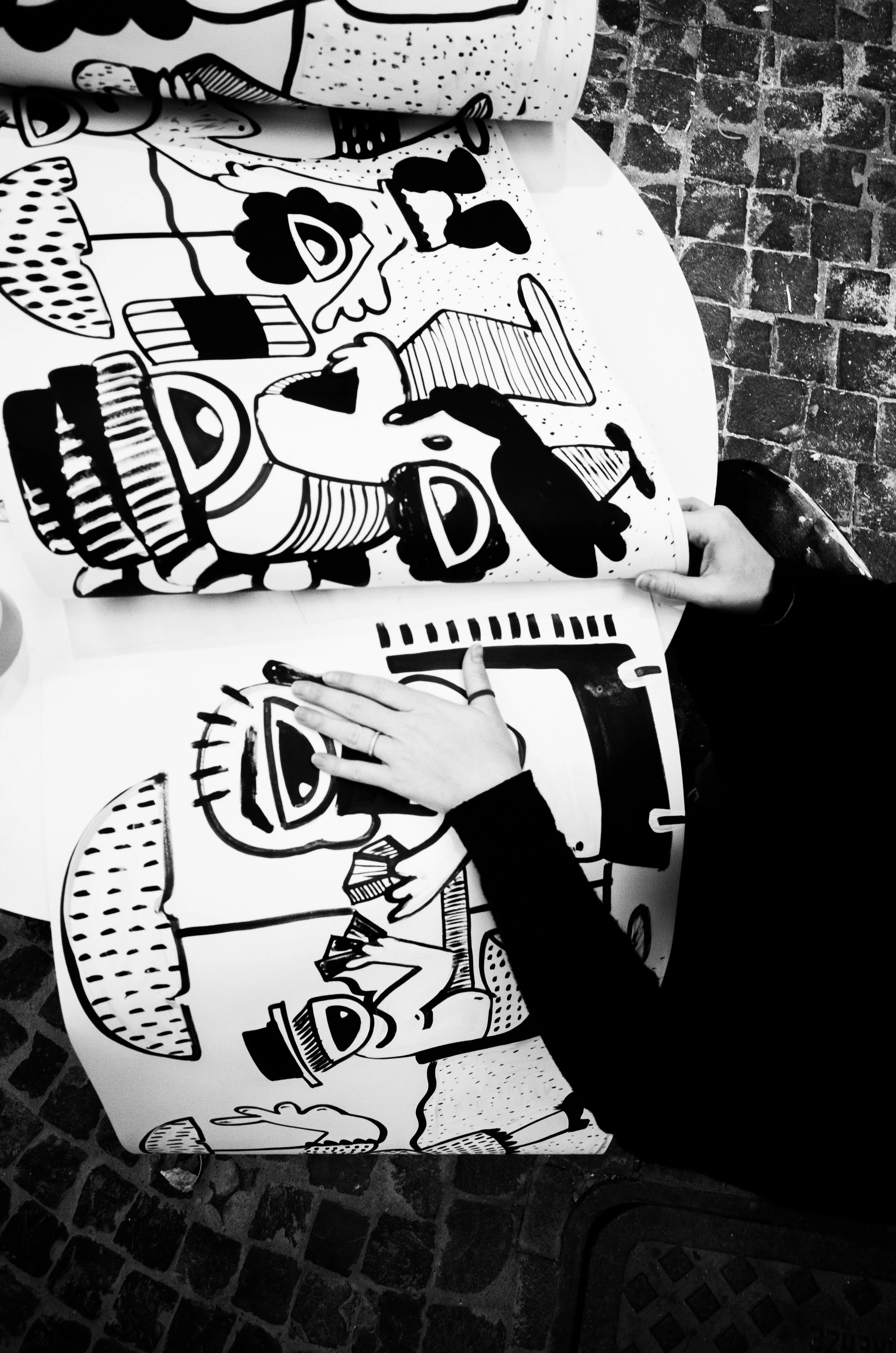 Two people, one young child and an adult, are coloring cartoon-style black and white drawings of people and objects on large sheets of paper on a round table, with a brick surface underneath.